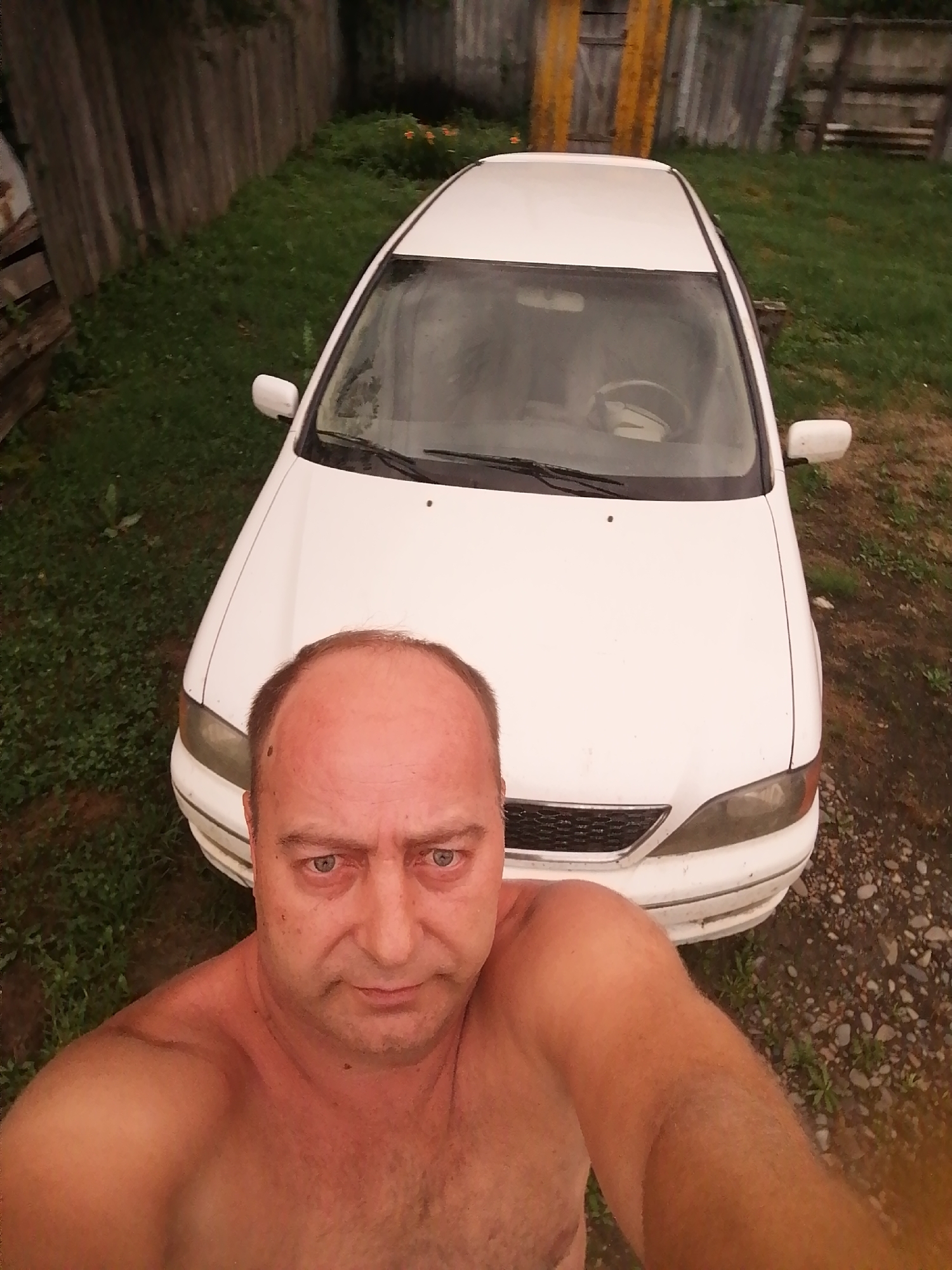 In Novopokrovka, Russia, a shirtless man stands in front of a white car on a gravel and grass driveway. The man is facing the camera, with his head and upper torso visible. The car is positioned behind him, taking up the majority of the frame. The background consists of a wooden fence and overgrown grass. The scene appears to be outdoors, possibly in a rural or suburban setting.