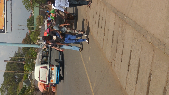 This image depicts a daytime street scene in Moshi, Tanzania. The scene includes a paved asphalt road with a faded yellow line marking the shoulder, adjacent to a concrete sidewalk featuring multiple linear drainage channels.
On the road, a white minibus (known locally as a Dala Dala) with dark horizontal stripes, a blue rear bumper, and yellow/red chevron reflective tape is either parked or moving slowly. Its rear license plate begins with "T 908". To the left of the road, several street vendor stalls with orange and red umbrellas are visible.
A group of four pedestrians is walking towards the right side of the frame, along the edge of the road, next to the sidewalk. From left to right:
1. A person wearing a dark top and light blue jeans, with a white cap, carrying a bag over the left shoulder.
2. A person wearing a black t-shirt with a light circular emblem, blue jeans, a black cap, and white sandals, carrying a dark blue bag.
3. A person wearing a green t-shirt, patterned trousers, and a pink cap.
4. Further right, a male individual wearing a light blue patterned shirt is partially visible.
To the right of the walking group, a male individual in a green shirt is pulling a wooden handcart filled with a dark, granular material.
In the background, there are various trees, residential and commercial buildings (some with green roofs), utility poles (one light blue, others brown), and a large billboard structure displaying orange and yellow graphics at the top right of the frame. The sky is partially visible and appears clear.