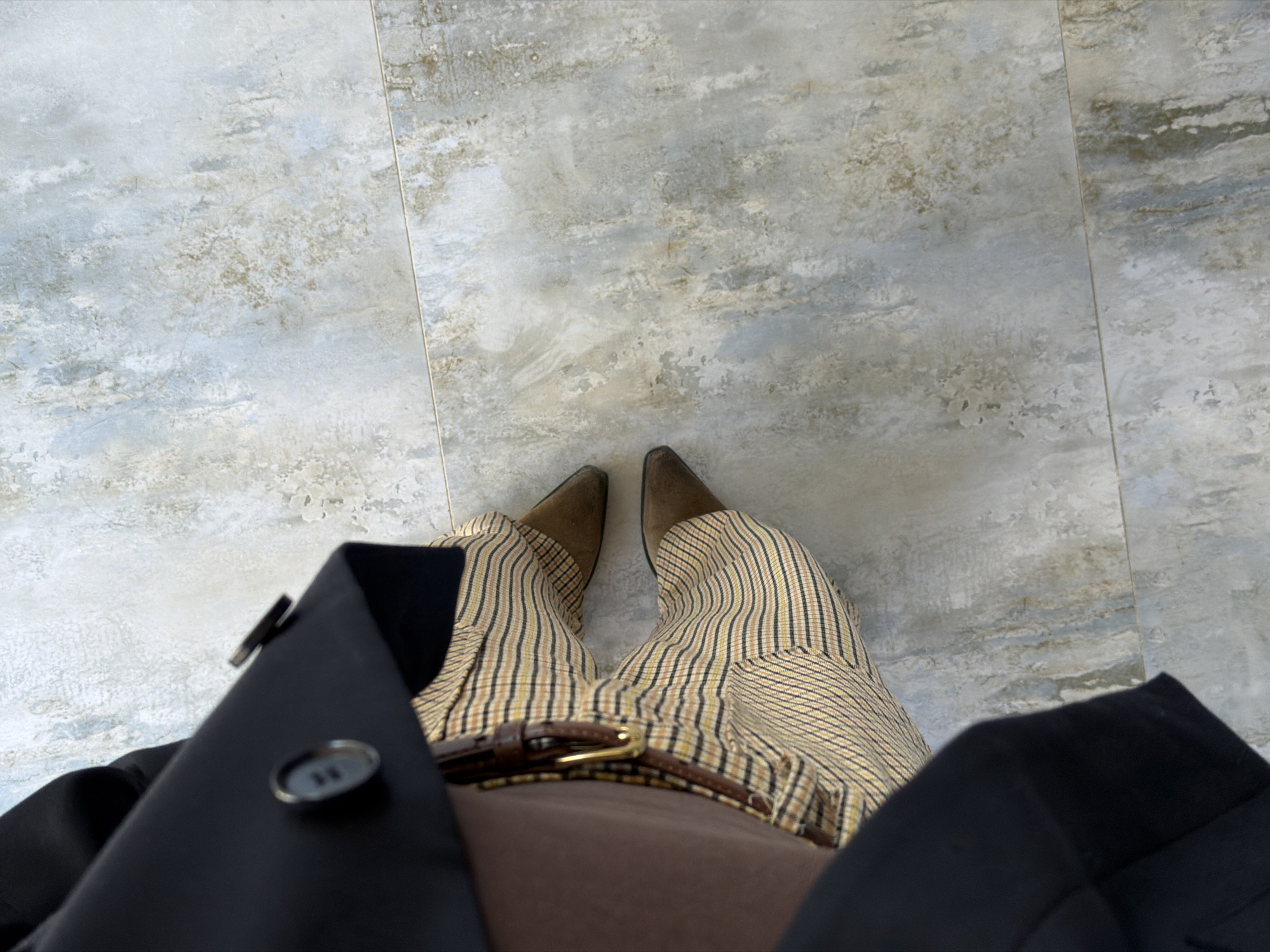 A top-down shot captures a person standing on light gray tiled flooring, possibly indoors. The person wears tan and brown plaid trousers with a brown belt cinched at the waist. A glimpse of a brown shirt is visible above the belt. Dark brown pointed-toe shoes are on the feet. Parts of a black coat are seen framing the lower portion of the image. The image does not provide sufficient information to determine if the location is in Yerevan, Armenia.
