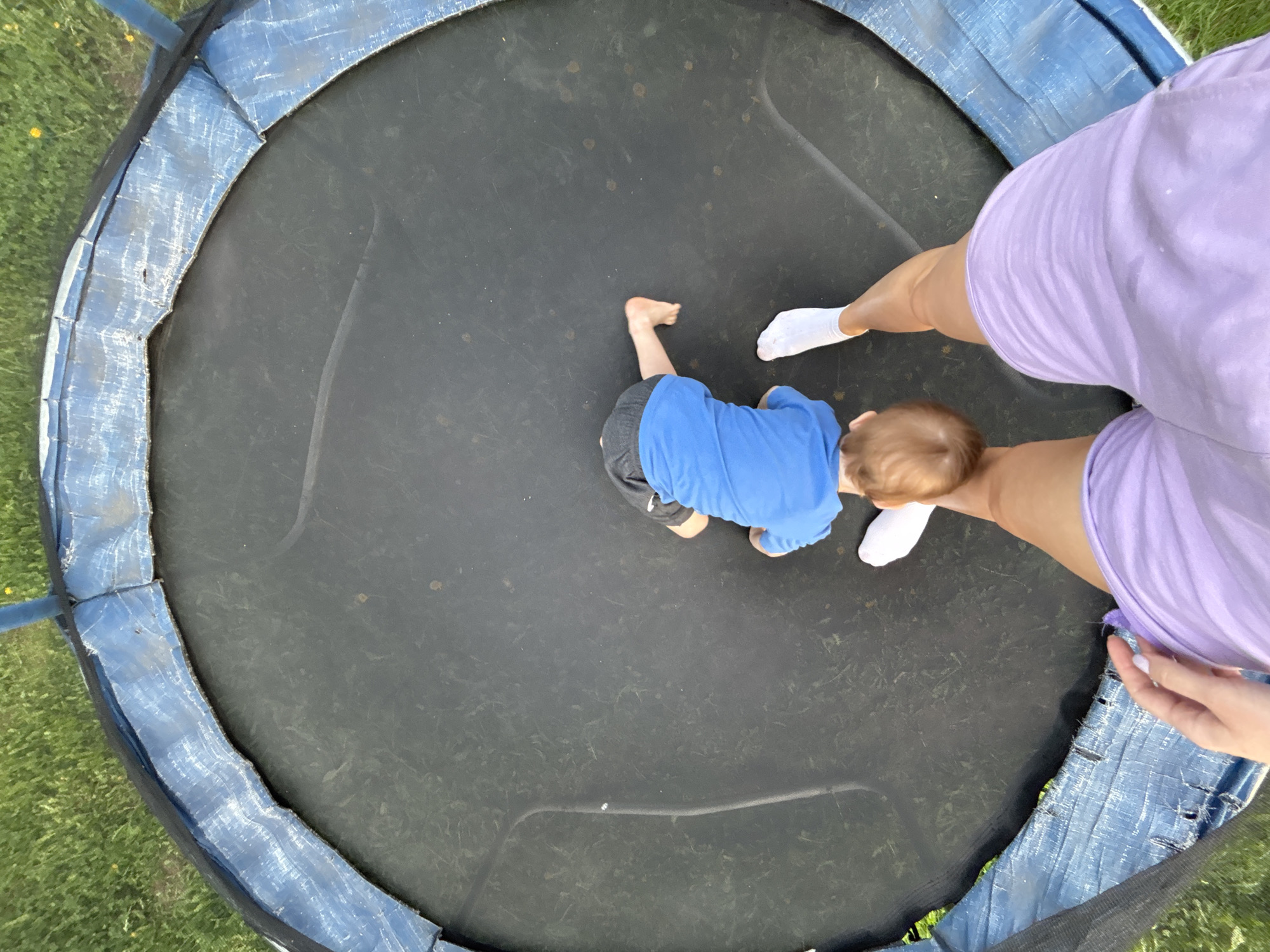 A high-angle shot depicts a child in a blue shirt and gray shorts crawling on a black trampoline. An adult wearing a lavender outfit and white socks is also on the trampoline. The trampoline has a blue padded safety edge. The background shows grass and a hint of trees, suggesting an outdoor setting. The location in Ферма, Russia is not verifiable from the image.