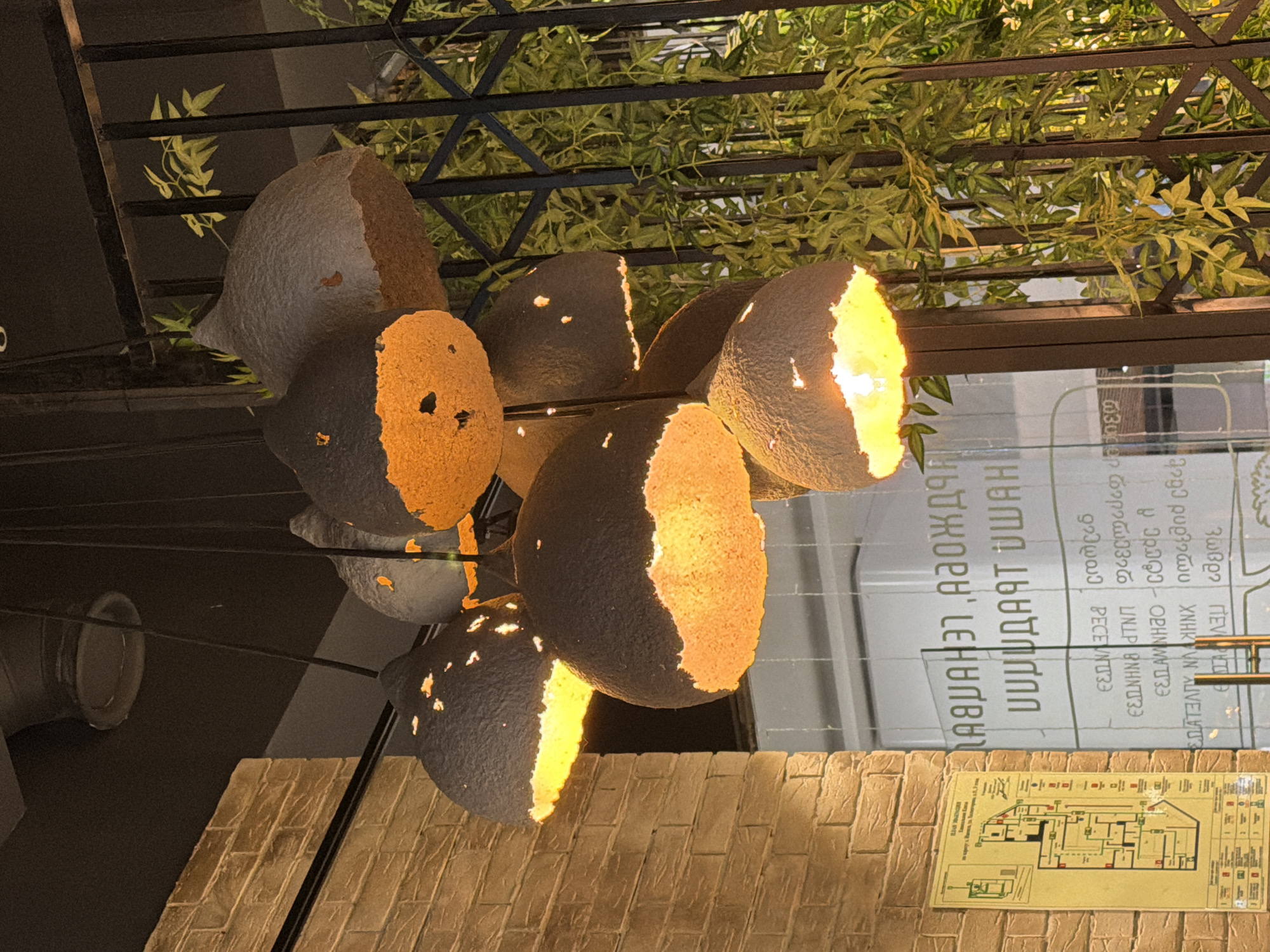 The image displays an interior or covered outdoor space, focusing on a unique light fixture and decorative elements. The central subject is a chandelier composed of approximately seven to ten roughly spherical or ovoid elements. These elements are dark gray or brown on the exterior and appear to be hollowed out or broken, revealing a rough, textured interior that glows with a warm orange-yellow light. The design evokes natural forms, possibly resembling abstract gourds or fragmented shells.
Above the light fixture, green, leafy vines are draped over a dark metal lattice or trellis structure, suggesting either artificial foliage or plants growing on a pergola. In the background, a large, reflective glass surface, possibly a window or partition, features prominent text in a non-Latin script that strongly resembles Georgian characters. The provided OCR output for this text is "ζωβρο Πεν", "2293 ρυρλητών ΧΝΗ UN ALVELUT", "EELAMINHO-Corde A", "RECENTЗЭ", "ПИРИДАЯТ ИШАН", and "ПАЏАНЭГ АГОЖДАН," which shows a mix of perceived Greek, Latin, and Cyrillic-like characters, indicating the challenging nature of recognizing the script from the reflective surface.
Below the glass surface, a textured wall with a reddish-brown brick pattern is visible. A small, detailed diagram, possibly a floor plan or area guide, is affixed to this brick wall. On the left, a grey ventilation duct or pipe is partially visible against a dark background.
The scene is illuminated by the warm artificial lighting of the fixture, suggesting an evening setting or an indoor environment where artificial light is dominant. The overall ambiance is warm and decorative, typical of a commercial establishment such as a restaurant or cafe. No people or specific activities are visible.