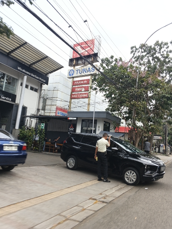 This is an outdoor daytime scene in front of an automotive dealership located at Jl. BKR No.17, Pelindung Hewan, Kec. Astanaanyar, Kota Bandung, Jawa Barat, Indonesia. A black multi-purpose vehicle (MPV) is positioned partially on a sidewalk and partially on the asphalt road in the foreground. A male security guard, wearing a light-colored polo shirt, dark trousers, and dark shoes, stands beside the driver's side of the black MPV, appearing to clean the window with his right hand while holding a yellow object in his left. A dark blue sedan is partially visible on the far left, moving or parked on the road.
The dealership complex features a building on the left with a dark canopy and signage for "Wicaksana Berlian Motor" and "Service." A taller, white building in the center displays a large "DAIHATSU" logo at its peak, below which a vertical sign reads "TUNAS" with sub-sections for "PENJUALAN" (Sales), "BENGKEL" (Workshop), and "SUKU CADANG" (Spare Parts). To the right of the main building, a small structure serves as a "POS SATPAM" (Security Post) with a "Mitsubishi Motors" logo and partial address numbers (175, 177, or 227) visible. Several individuals are seated on benches under an awning near the dealership entrance. A large tree with green foliage and pink blossoms stands on the right side of the frame. Numerous black utility cables crisscross overhead against an overcast sky. The sidewalk features tactile paving strips.