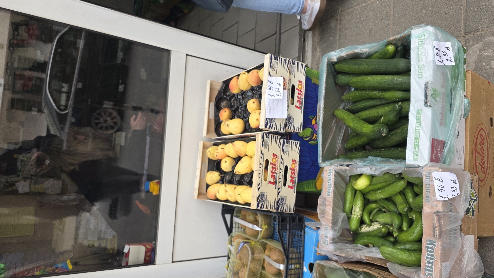 The image captures an outdoor street-level scene in Blagoevgrad, Bulgaria, featuring a display of fresh produce in front of a shop. The main subjects are various fruits and vegetables arranged in wooden and cardboard crates.
Two wooden crates are stacked, holding yellow-orange, pear-shaped fruits, possibly quince or a type of pear, resting on black molded trays. These crates are labeled "Latsios PRODUCTS". Below these, a black plastic crate contains packaged kiwi fruits.
To the right, two cardboard boxes are filled with green cucumbers. The top box, labeled "Sidonia Sun" and "CUCUMBERS of PSARI", contains larger cucumbers. The bottom box, marked "Κρήτης" (Crete) and "Κηπευτικά" (vegetables), holds smaller, gherkin-like cucumbers. Another partially visible cardboard box displays "delore".
Price tags are affixed to several crates. The fruit is priced at "1.90 €" with a weight indication "3.72 k" (likely kg). The large cucumbers are "1.20 €" for "2.35 k", and the smaller cucumbers are "1.50 €" for "2.93 k". These prices and Greek labeling suggest imported produce, common in Blagoevgrad due to its proximity to Greece.
In the background, a large glass window reflects parts of a street, including the wheel and side of a car, and indistinct figures, one showing a person's hand. The lower legs of a person wearing jeans and white sneakers are visible at the top edge of the frame, indicating someone standing near the display. The lighting suggests daytime, likely overcast, with no harsh shadows. The overall scene depicts a small, open-air produce stall or a grocery store's external market display.