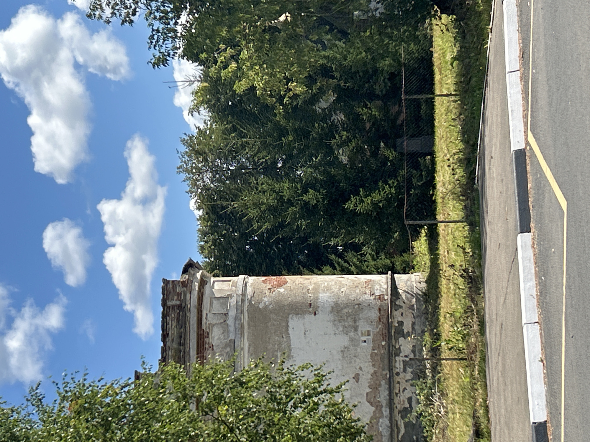 An abandoned, old, round building with peeling plaster and exposed brickwork stands partially obscured by lush green trees. The building appears to be in disrepair, with its top section crumbled. A chain-link fence runs along the front of the property, separating it from a paved road with a yellow line. The sky above is a clear blue, dotted with fluffy white clouds. The overall scene suggests an overgrown and neglected area, possibly on the outskirts of Kalitsino, Russia, given the context provided.