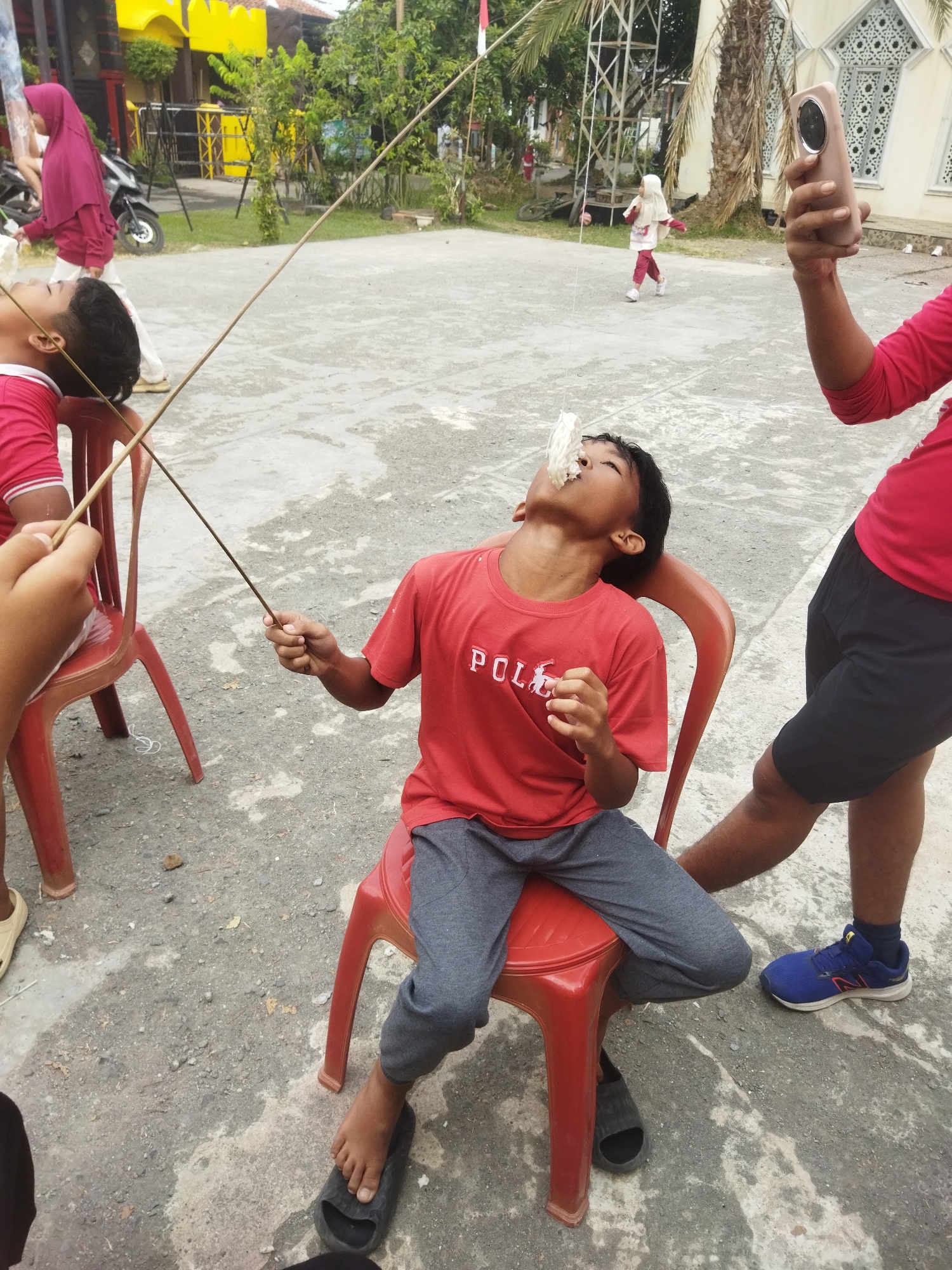 A group of children are participating in a game of eating crackers suspended from strings in an outdoor setting, possibly a street or yard. The main subject is a young boy sitting on a red plastic chair, his head tilted back as he attempts to eat a cracker held by a string in front of his face. He is wearing a red t-shirt with "POLO" written on it and grey pants. Another child, to the left, is partially visible and also appears to be participating in the game. In the background, a young girl in a hijab is walking, and another child in a white hijab and red pants is running. An adult's arm, wearing a red shirt, is visible on the right, holding a smartphone, suggesting someone is recording the event. The ground is paved with concrete, and there are buildings and trees in the background, indicative of an urban or suburban environment. The scene suggests a community event or a celebration in Banjar, Indonesia, given the architectural style of the building in the background which resembles a mosque, common in Indonesia.