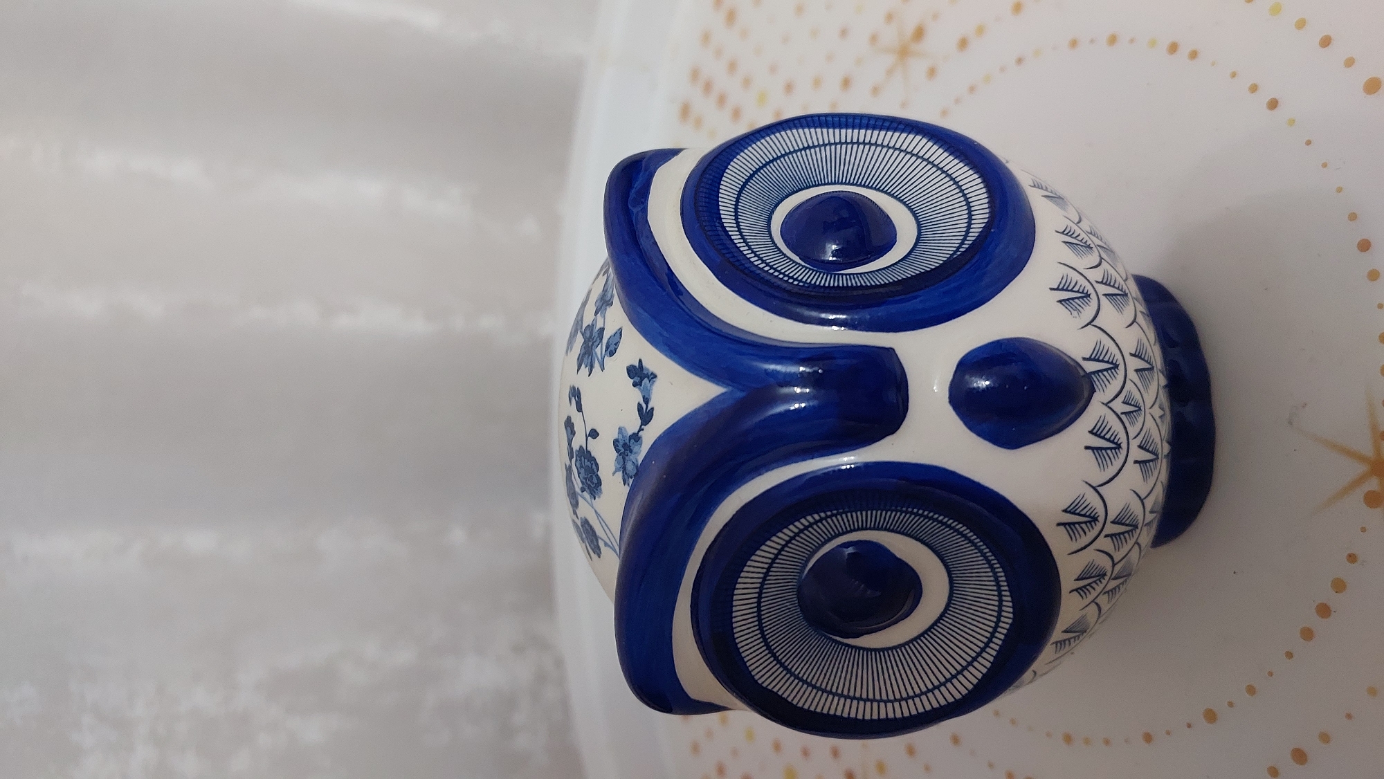 A close-up, head-on shot showcases a white ceramic owl figurine with blue painted detailing. The owl has large, concentric blue circles for eyes with intricate line work, a small blue beak, and blue wing-like shapes above the eyes. The top of its head is adorned with small blue floral patterns. The owl sits on a white, circular surface with gold starbursts and scattered gold dots, possibly a decorative tray. In the background, a white, textured curtain is visible. The image is taken indoors, with soft lighting. The location is the city of Novocheboksarsk, Russia, within a residential interior.