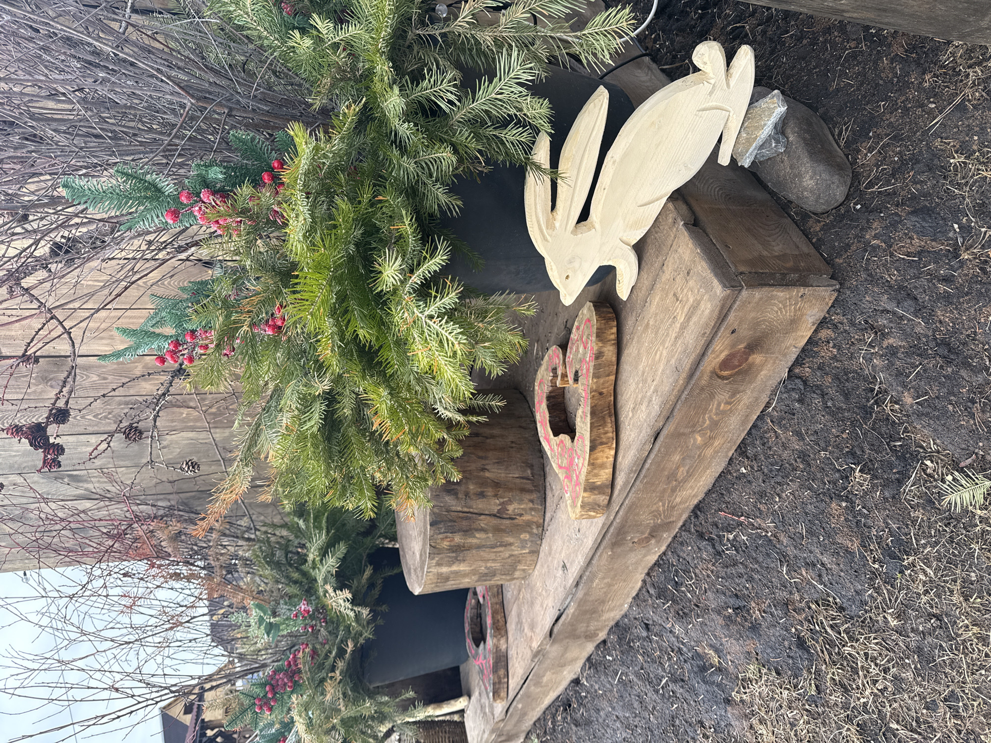 A static outdoor decorative display is situated in Tyulyuk, Russia. The scene features a weathered wooden plank platform resting on dark, damp soil interspersed with dry grass and sparse fallen detritus. On the platform, from left to right, are two dark potted evergreen conifers, likely fir or spruce, adorned with clusters of artificial red berries and interspersed bare twigs. Between these potted plants, a substantial, vertically oriented section of a wooden log with bark remnants stands, accompanied by two irregularly shaped, flat wooden cutouts, each displaying faded red painted patterns. To the right, a light-toned, flat wooden cutout depicting a rabbit rests partially on the platform's edge and partially on two gray rocks positioned on the soil below. In the background, behind the display, a weathered vertical wooden fence or wall is visible, backed by dense bare deciduous branches and the upper sections of distant buildings with dark roofs, all under an overcast sky. No individuals are present in the frame.