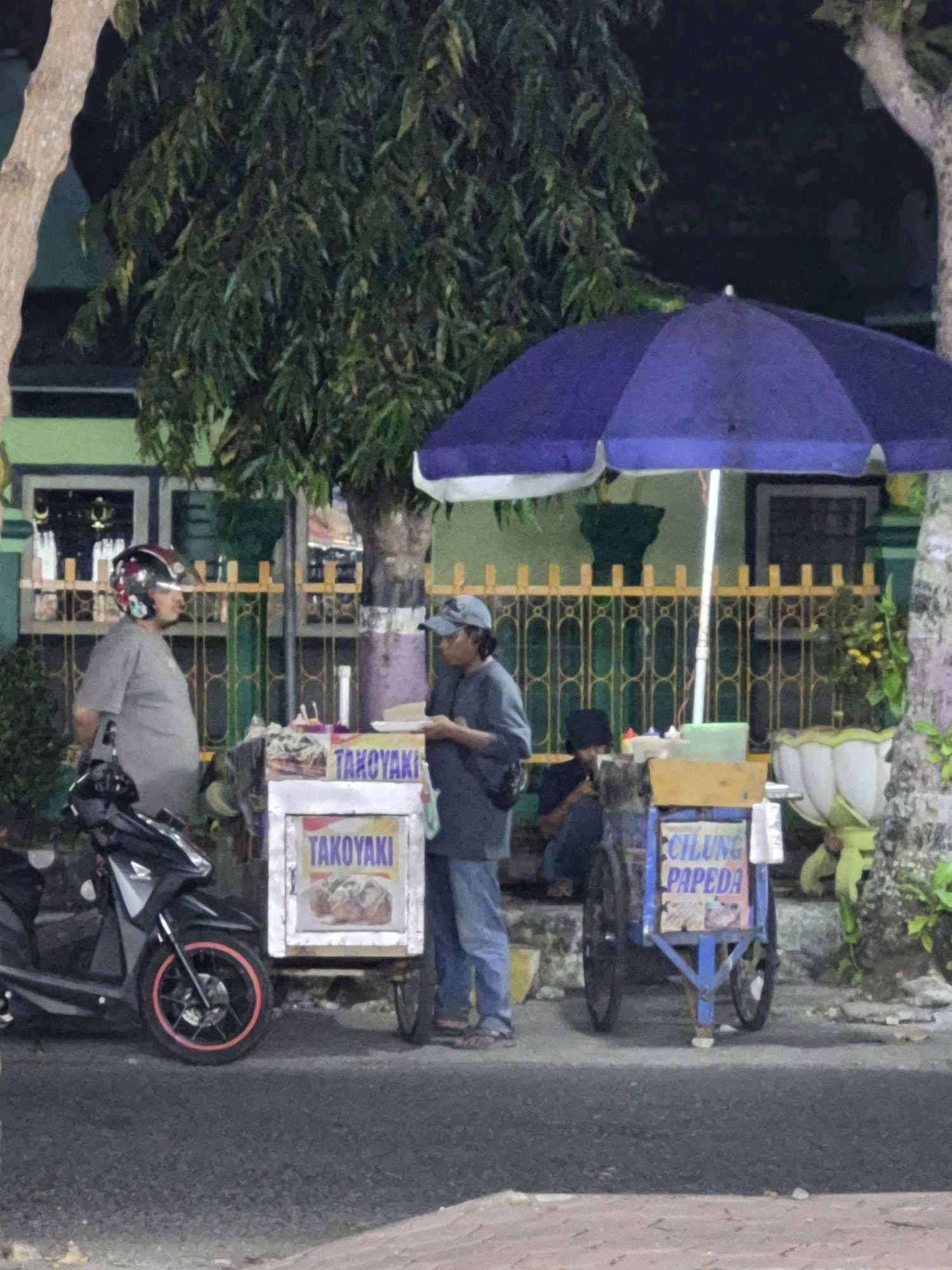 The image depicts a street scene at night in Purworejo, Indonesia, featuring two food carts and a motorcycle. A person wearing a gray t-shirt and a helmet stands next to a black motorcycle with a red rim on the front wheel. Adjacent to the motorcycle is a food cart with a sign that reads "TAKOKYAKI" and displays an image of food. To the right of this cart, another person in a blue shirt and cap is interacting with a second food cart. This cart is blue and has a sign that reads "CILUNG PAPEDA". A large purple umbrella shades the area around the second food cart. In the background, a green fence with yellow ornate details separates the street from a building. Trees and plants are visible, some of which are illuminated by artificial light. The overall setting suggests a common street food vending area in Purworejo.