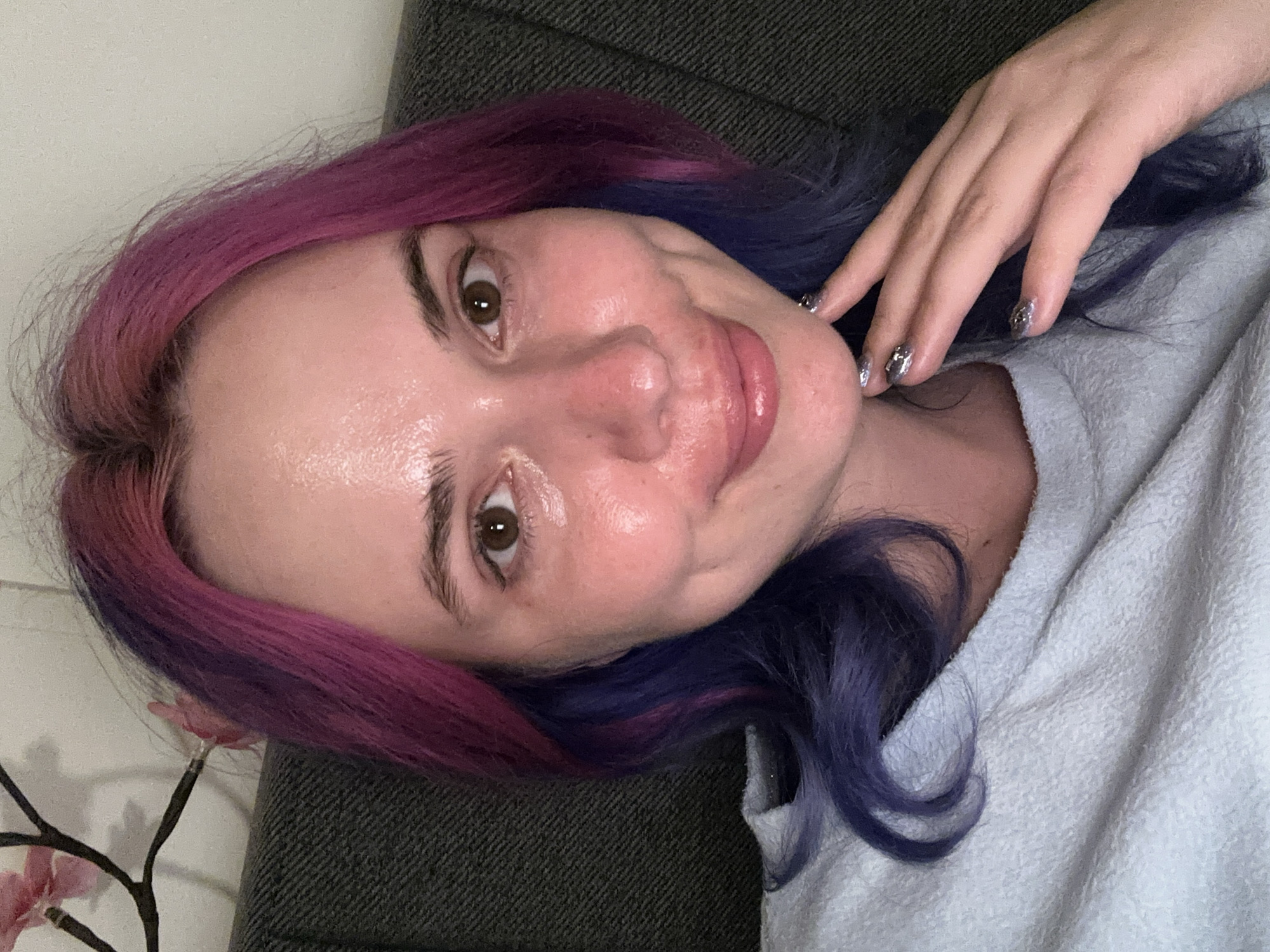 A person with purple and blue hair is lying on a gray surface, possibly a couch. The person is facing the camera with their right hand near their face. The person has brown eyes. Their skin appears shiny. The person is wearing a light-colored shirt. Behind the person is a white wall with a floral decoration. The location is not possible to determine from the image.