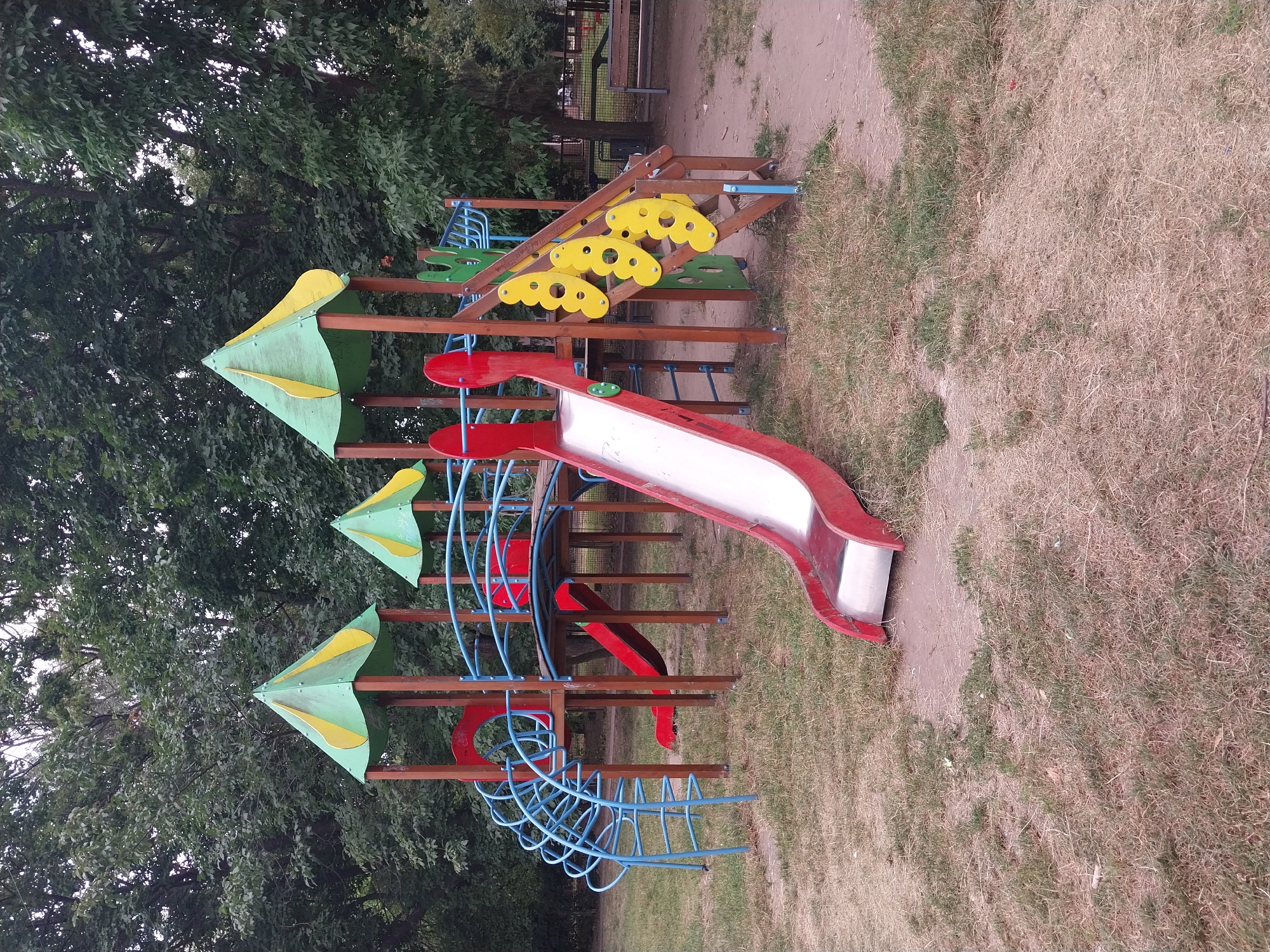 This image depicts a children's playground in Doslidne, Ukraine, situated amidst a backdrop of lush green trees. The playground features a multi-level structure constructed primarily of wood and metal. Prominently, there is a red and silver metal slide with a red frame, and a blue metal climbing structure resembling a spiral staircase. Another slide, a red one, is partially visible. The playground's upper levels are adorned with green and yellow conical roofs. Wooden posts support the structure, and yellow, butterfly-shaped cutouts are integrated into the stairs on one side. The ground surrounding the playground is covered with dry grass and some bare patches of earth. In the background, a park bench is visible, and a fence runs along the edge of the playground area. The scene is static, with no people or animals present.