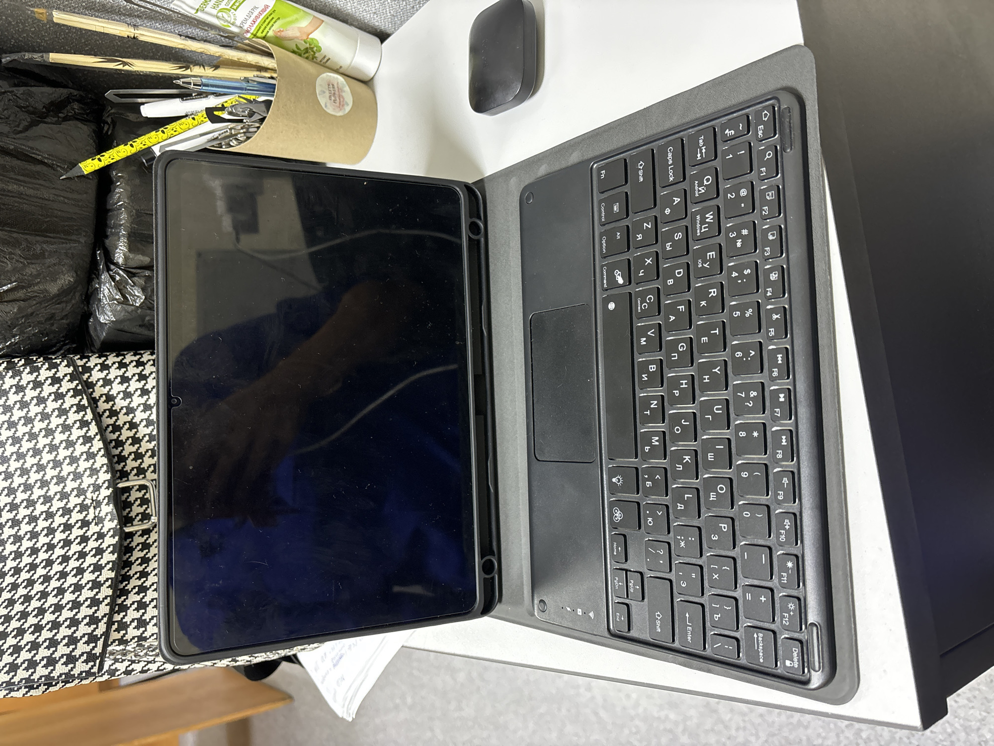 The image shows a partially opened tablet with an attached keyboard on a table. A black wireless mouse is placed above the tablet. Next to the tablet are a pencil holder with pens, pencils and a tube of hand cream with Russian text. A houndstooth patterned bag is seen on the left, and a black trash bag is visible near the pencil holder. The keyboard displays both Latin and Cyrillic alphabets.
While the Russian text on the cream tube suggests the location may be in Russia, there is no explicit evidence in the image to confirm the specific city of Chaikovsky.