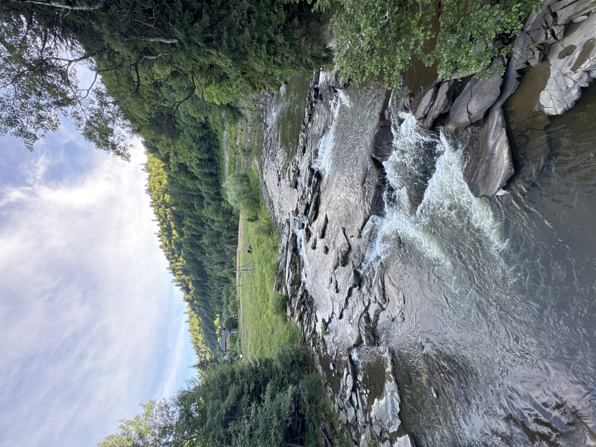 The image shows a rocky river flowing through a lush, green valley. The riverbed is filled with numerous large, flat stones, creating small rapids and waterfalls as the water cascades over them. The surrounding landscape is dominated by dense forests that cover the hillsides, with a patch of green meadow visible in the distance, featuring a few buildings and some grazing animals. The sky above is partly cloudy, with streaks of white clouds against a light blue backdrop, suggesting it is daytime. The overall impression is a serene, natural environment in a rural setting, likely in the Carpathian Mountains near Tatariv, Ukraine, given the characteristic landscape and architecture.