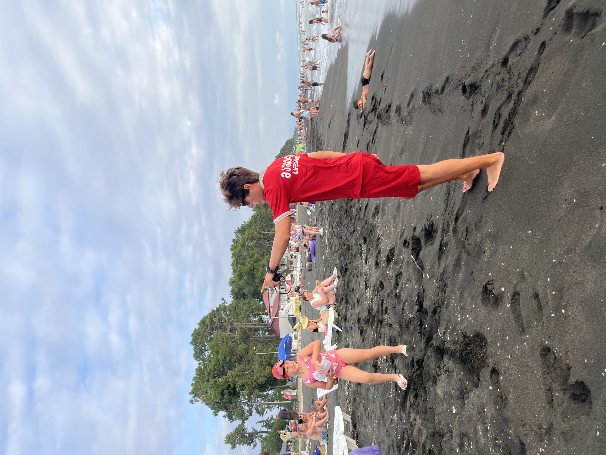 A lifeguard in a red shirt and shorts is standing on a black sand beach in Ureki, Georgia, pointing towards the water. A young girl in a pink swimsuit and goggles is standing to his left, holding a clear bottle. In the background, numerous beachgoers are visible, some wading in the shallow water, others relaxing on lounge chairs or towels. The sky is overcast with scattered clouds, and a line of green trees borders the beach. The overall scene depicts a lively summer day at the seaside resort.