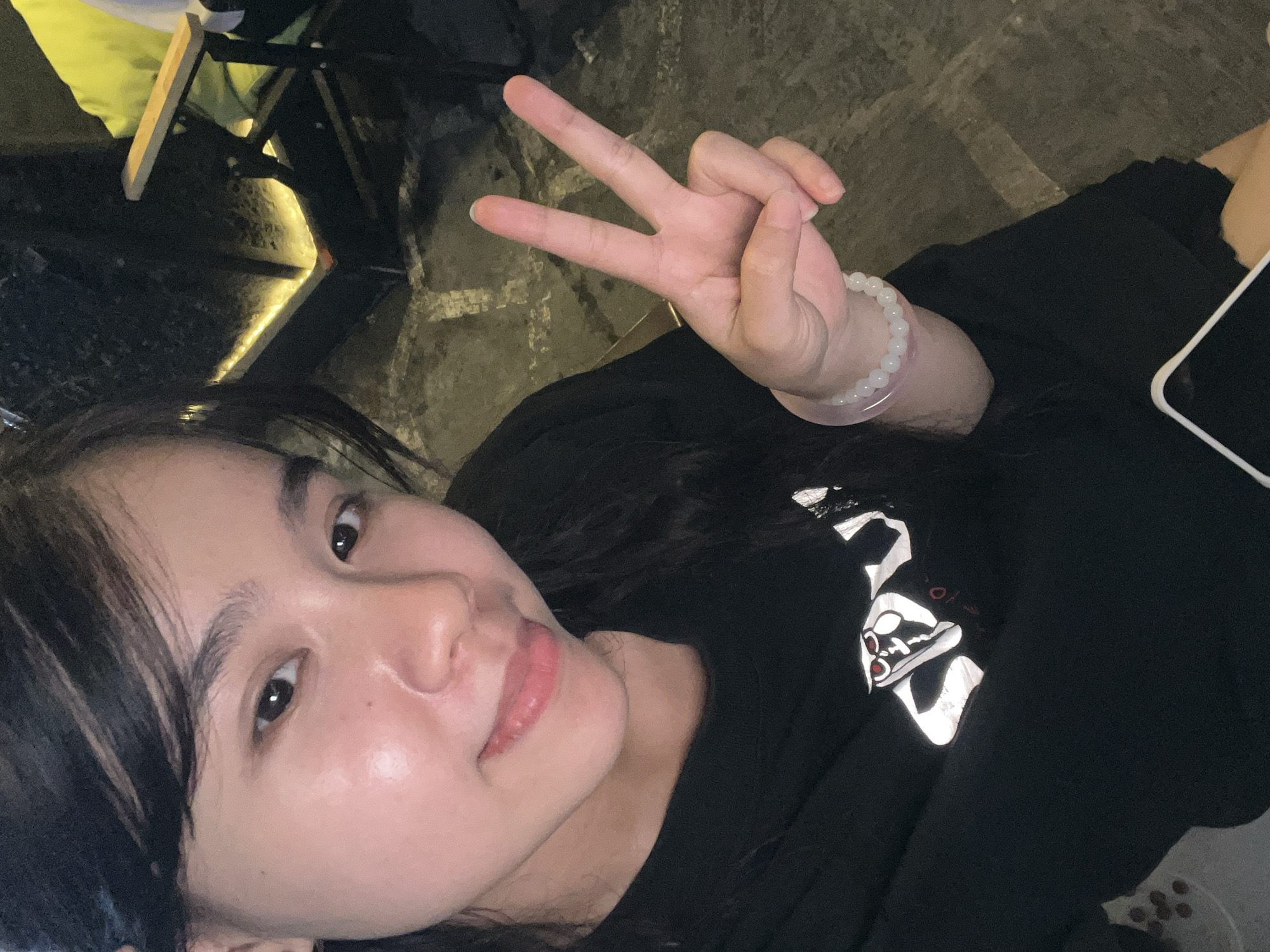 A person with dark hair and fair skin is positioned in the lower-left quadrant of the frame, angled upwards towards the camera. They are giving a "peace" or "victory" sign with their right hand. The hand features a white beaded bracelet. The person is wearing a black top with a reflective white design visible on the front. Part of a white-cased mobile phone is visible. To the left of the person, is a cafe table with a reflective top and chair. The background consists of a dark, textured surface, possibly a floor. While it is not possible to ascertain if this image was taken in Đà Nẵng, Vietnam.