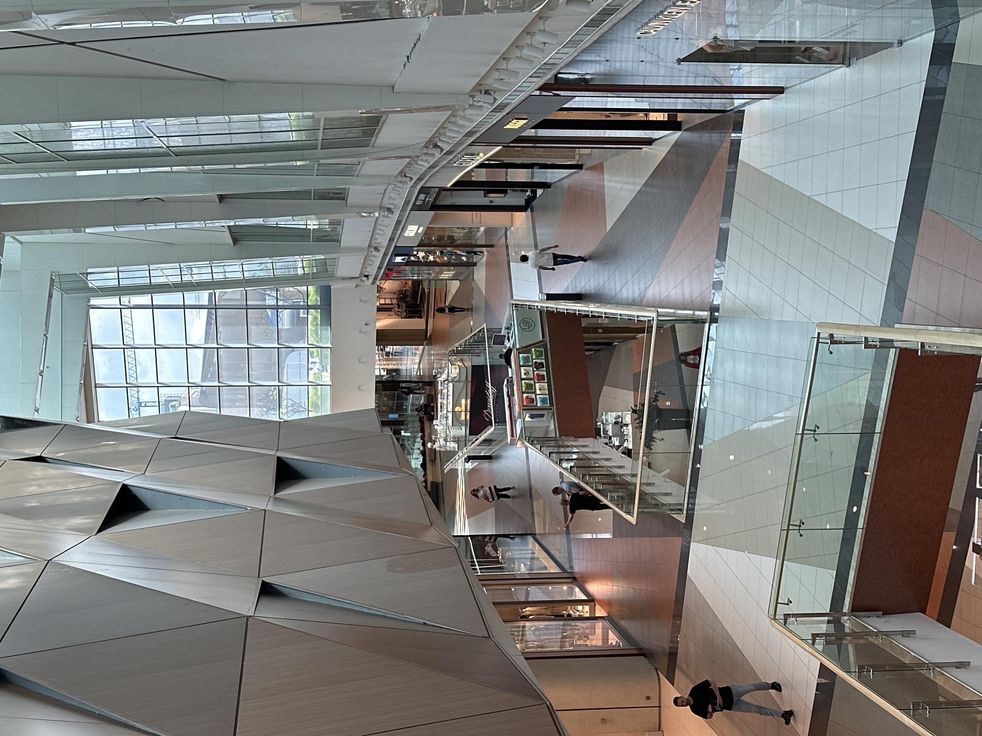 A high-angle shot captures the interior of the Paradise Center shopping mall in Sofia, Bulgaria. The scene reveals multiple levels, walkways, and shops. People are dispersed throughout the mall, some walking and some browsing in the shops. The architectural design is modern with geometric patterns, glass railings, and skylights. A triangular, geometric structure dominates the left foreground. A large window with a grid pattern is seen in the left midground.