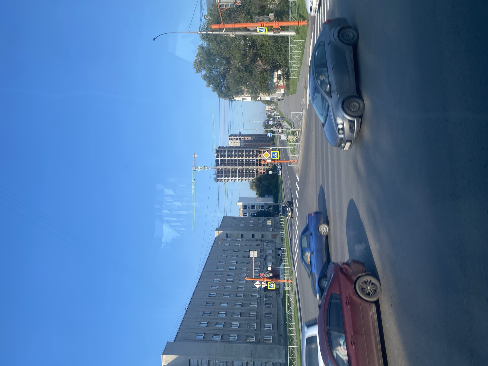 The image captures an urban street scene in Kemerovo, Russia, likely during the daytime due to the clear blue sky and natural lighting. The perspective suggests the photo was taken from a moving vehicle.
In the foreground, several cars are in motion. A dark red sedan is on the left, emitting a small puff of white exhaust. To its right and slightly behind, a blue sedan is traveling in the same direction. Further to the right, a dark gray sedan is crossing an intersection.
The midground features a multi-lane asphalt road with pedestrian crossings. Traffic signals and yellow pedestrian crossing signs are visible on orange poles at the intersection. On the left side of the road, a long, multi-story concrete apartment building with numerous windows stretches across the frame. On the right side, trees and a sidewalk are present, with another smaller building visible further down the street.
In the background, the cityscape is characterized by the construction of several high-rise buildings, with cranes actively present, indicating ongoing development. Power lines crisscross the sky above the scene. The overall environment is that of a busy urban area undergoing construction.