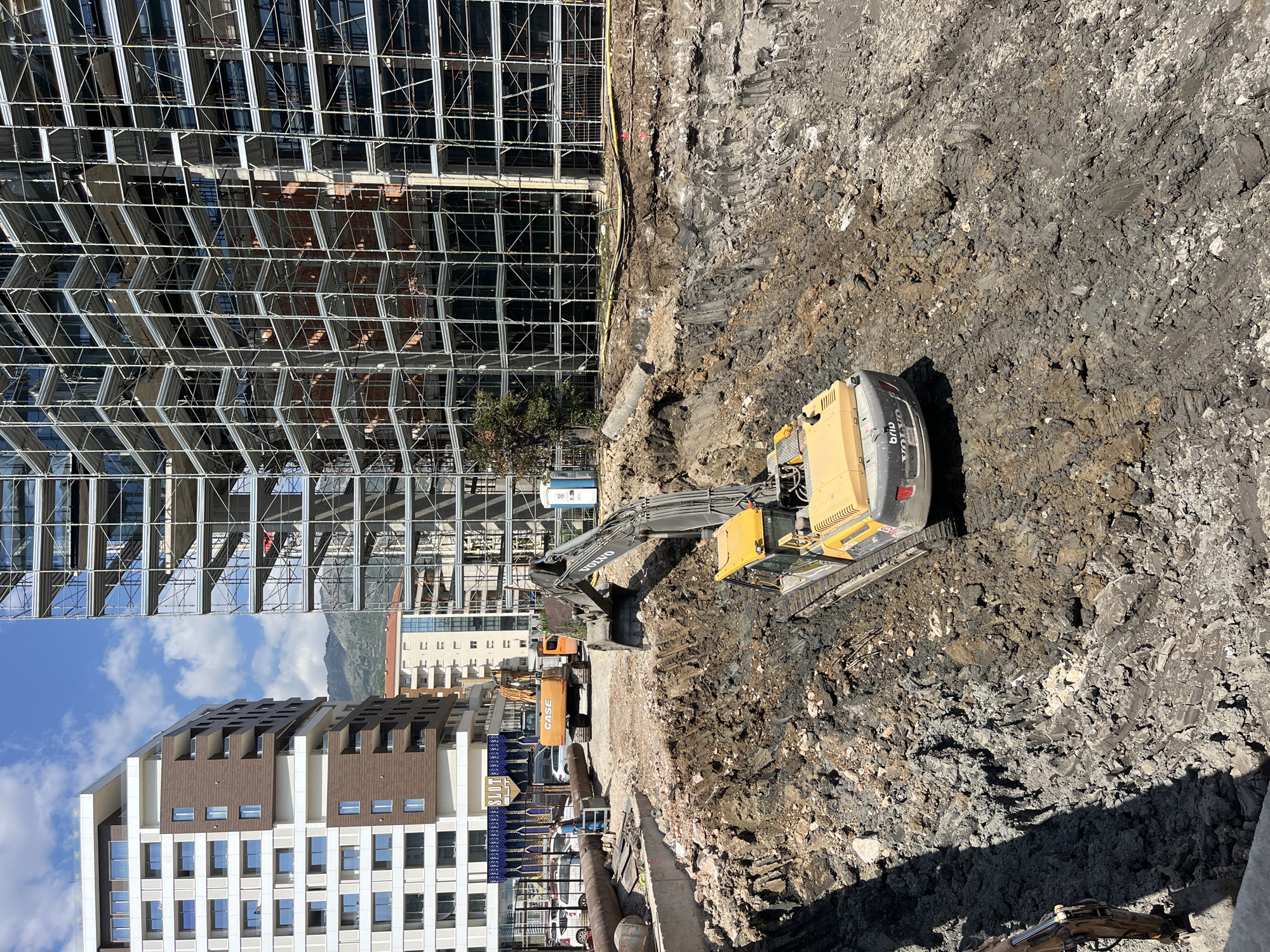 The image shows a construction site in Budva, Montenegro. A tall building with scaffolding occupies the left side of the frame. An excavator is digging in the foreground, surrounded by excavated earth and rocks. Another excavator and construction materials are visible in the background. A completed building stands behind the construction site. Mountains are visible in the distance, under a blue sky with clouds. A portable toilet is on site near the scaffolding.