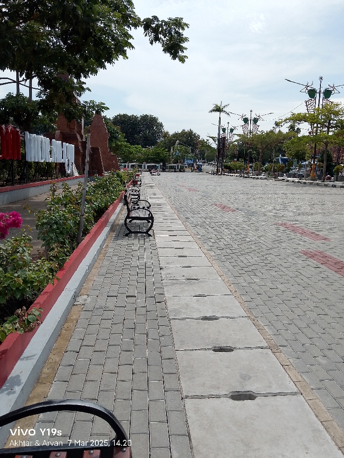 The image shows a paved walkway with concrete benches running alongside the edge of the walkway. The walkway is made of gray bricks with red and white painted lines running along the edges. There is a gray concrete sidewalk bordering the walkway. There are trees in the background and a sign in the distance that reads "INDONESIA." The location is in Indramayu, Indonesia, at coordinates -6.326 latitude and 108.323 longitude. There are no people in the scene.