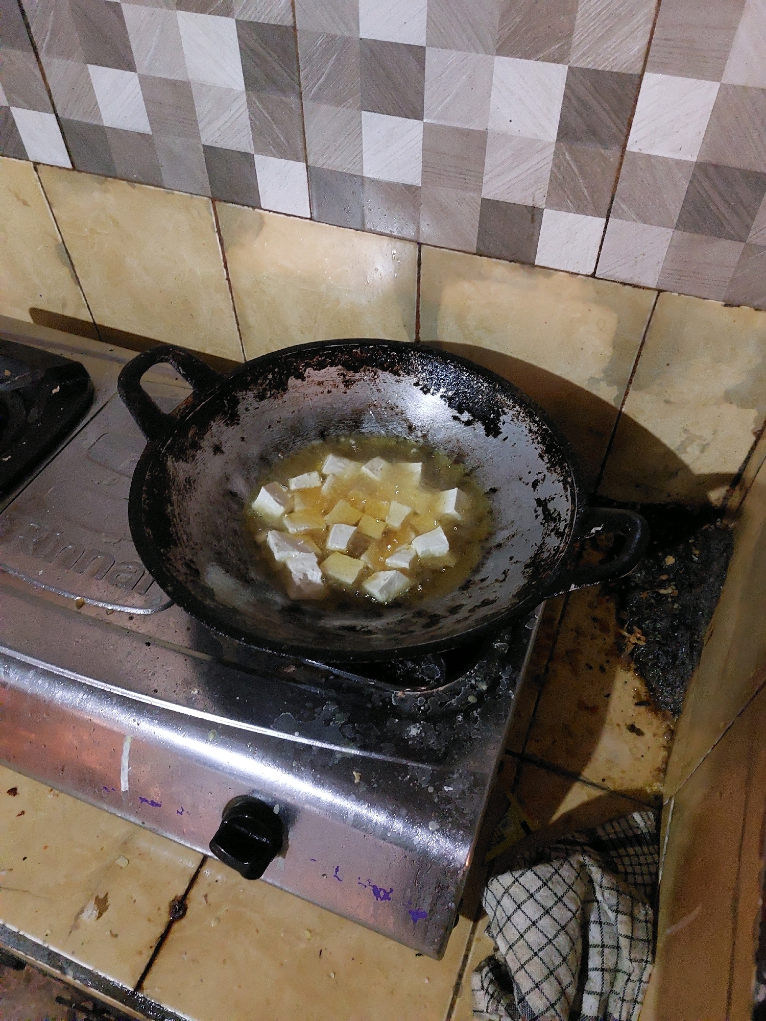 A close-up, overhead view of a black, well-used frying pan filled with cubed tofu sizzling in hot oil sits on a stainless steel gas stove. The pan has two dark handles, one on each side, and the oil is bubbling around the tofu. The stove has a silver surface with a black knob on the front. The background features a tiled wall with a pattern of beige and gray squares. To the right of the stove, there is a small pile of dark debris, and a folded checkered cloth is visible at the bottom right corner. The overall scene depicts a cooking preparation in a kitchen, likely in Sarangan, Indonesia.