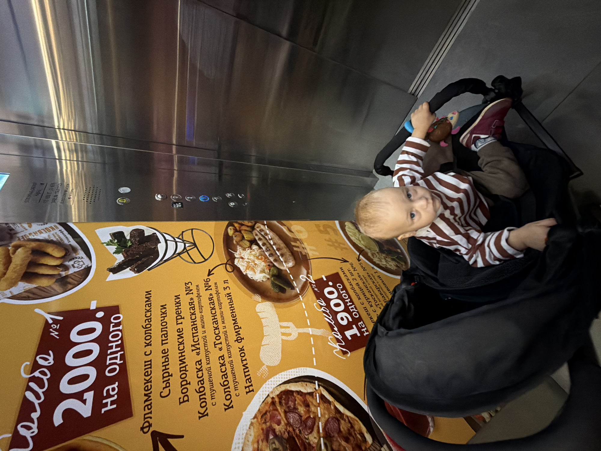 The image depicts a young child sitting in a stroller in an elevator. The child is wearing a striped shirt and brown pants. The stroller is black and positioned in front of a large yellow advertisement featuring food items and prices. The advertisement lists "Кольбо 2000. На одного" which translates to "Kolbo 2000. Per person." and "Кольбо 1900. На одного" which translates to "Kolbo 1900. Per person." Some of the food items mentioned include "Фламекеш с колбасками" (Flamekesh with sausages), "Сырные палочки" (Cheese sticks), "Бородинские гренки" (Borodino croutons), and various types of sausage dishes like "Колбаска «Испанская»" (Spanish sausage), "Колбаска «Тосканская»" (Tuscan sausage), and "Баварская" (Bavarian). The elevator doors are visible behind the stroller and advertisement. The elevator interior is metallic and reflects light. The context suggests the location is a public place, potentially a shopping mall or a restaurant, in Khimki, Russia, given the Cyrillic text on the advertisement.