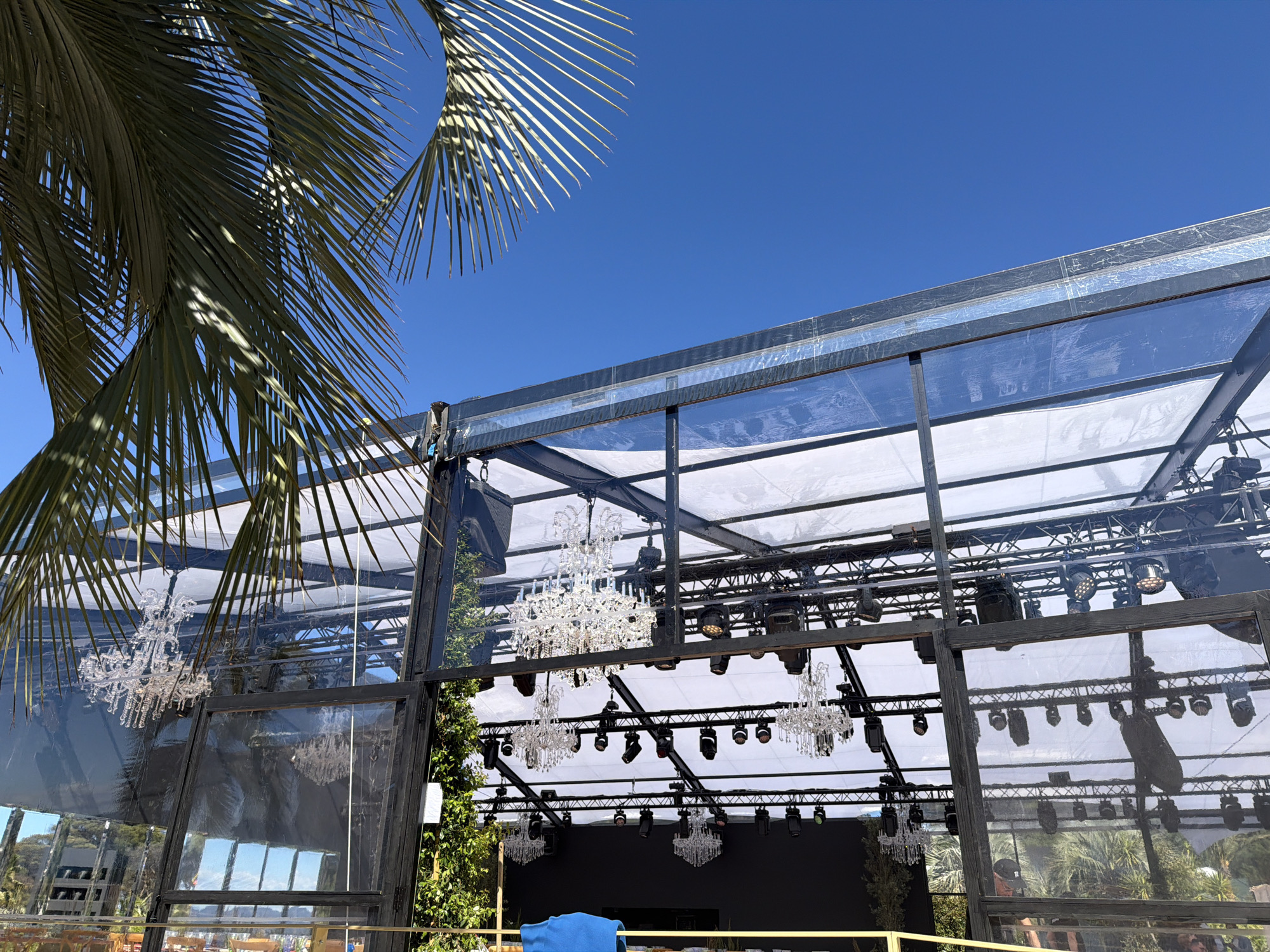This photograph, taken in Saint-Tropez, France, depicts a venue with a stage set up under a large, open structure. The structure is constructed from steel and clear panels and is suspended with several crystal chandeliers. There are numerous stage lights and rigging visible above the stage area. The venue features a palm tree, partially obscuring the view of the clear blue sky.