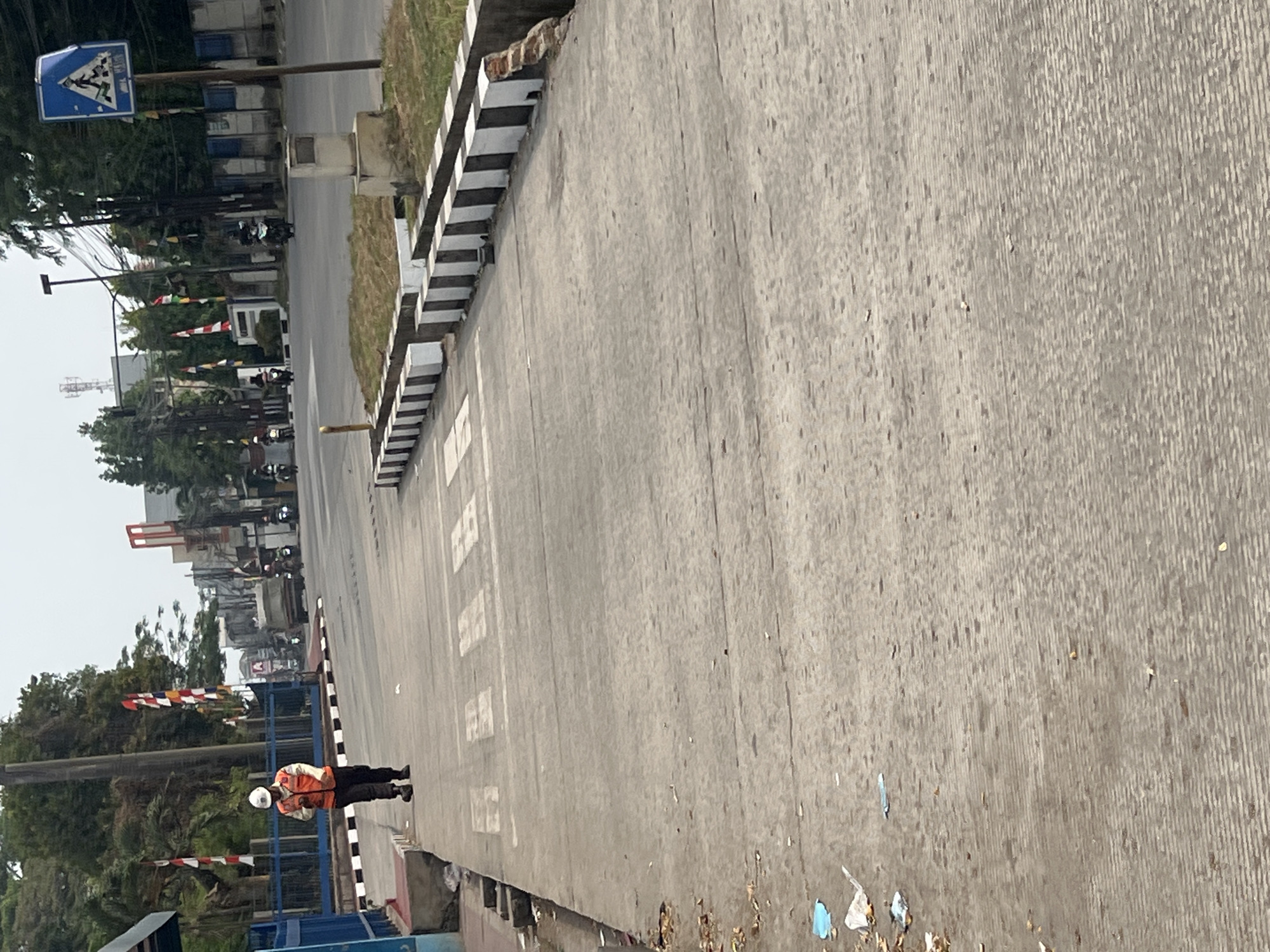 In Citeureup, Indonesia, a person wearing a white hard hat and an orange vest is walking on a pedestrian walkway next to a road. The walkway has a zebra crossing painted on it. The road is busy with traffic, including motorcycles and a truck. There are trees and buildings in the background, as well as flags. The sky is overcast. There is litter on the walkway and on the side of the road.