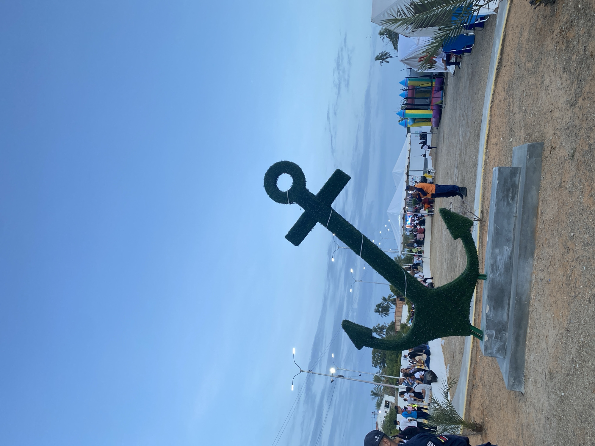 The image shows a large green anchor sculpture made of greenery, likely artificial, standing on a concrete base in an outdoor setting in Porlamar, Venezuela. The sky is clear blue, transitioning to a lighter hue towards the horizon. In the background, there are white tents, a colorful inflatable structure, and people gathered, suggesting an event or fair. Streetlights line the area. The ground is sandy or gravelly. The image is taken from a low angle, with the anchor dominating the foreground.