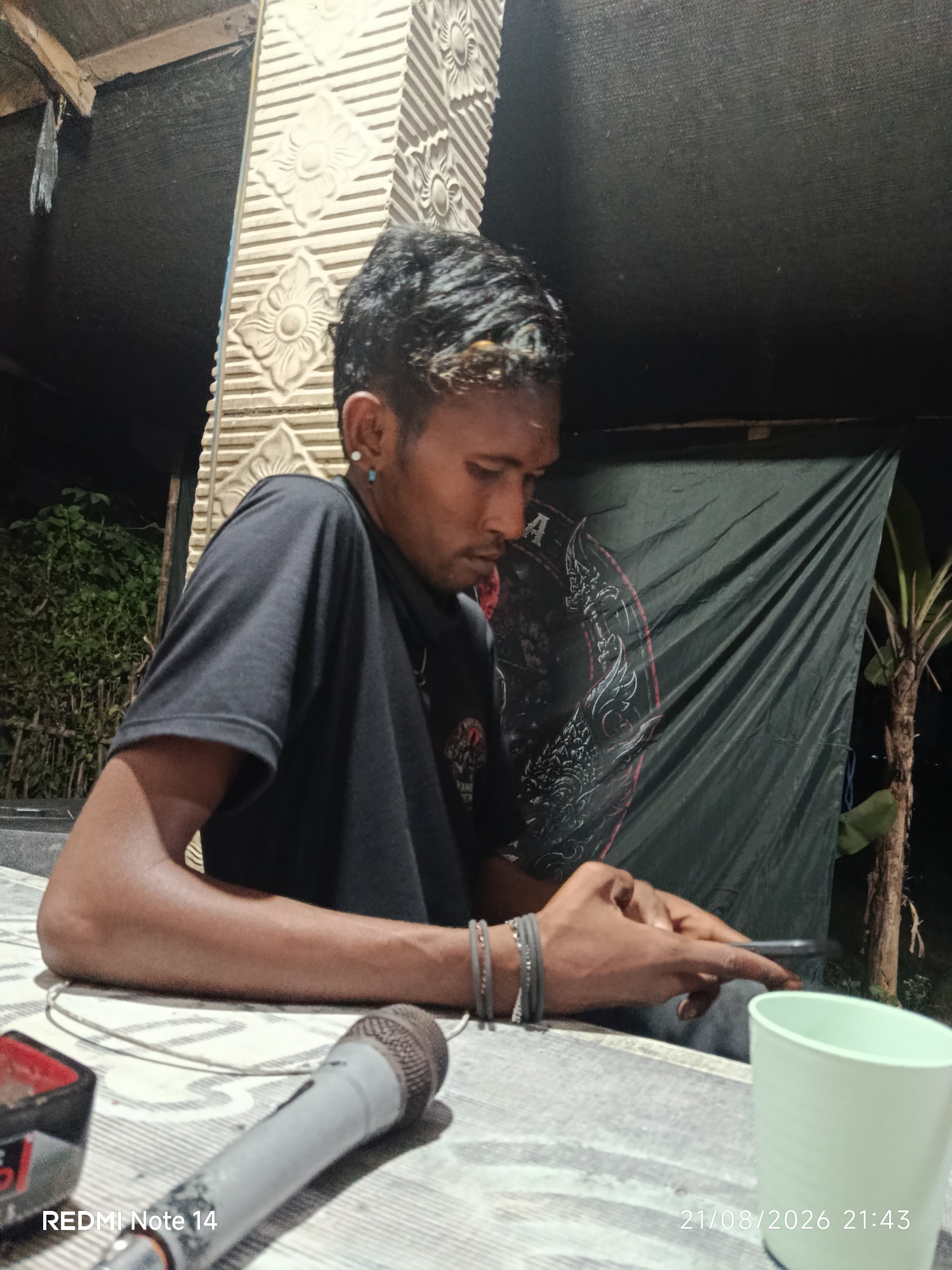A young man with dark, styled hair and a blue earring is seated at a table, looking down at his smartphone. He is wearing a black t-shirt and several beaded bracelets on his left wrist. In front of him on the table are a microphone, a red and black object, and a light green cup. Behind him, a dark tarp with a circular, intricate design hangs on the wall. To the left of the man, there are green plants and foliage. The scene appears to be outdoors at night, possibly in a rural or semi-urban area, as suggested by the presence of plants and the natural lighting. The timestamp in the lower right corner indicates the image was taken on August 21, 2026, at 21:43. The location is identified as Panggungrejo, Indonesia.