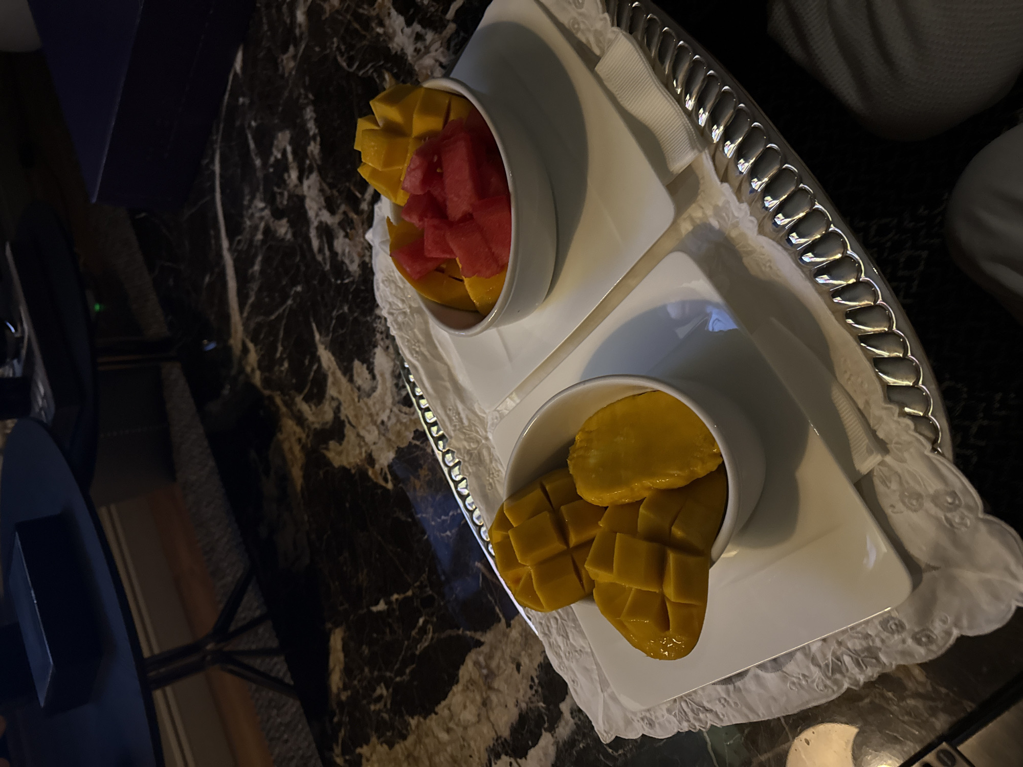 On a marble table in Mexico City, Mexico, a silver tray holds two white bowls. One contains diced mango and watermelon. The other contains sliced mango. The tray is lined with white cloth.