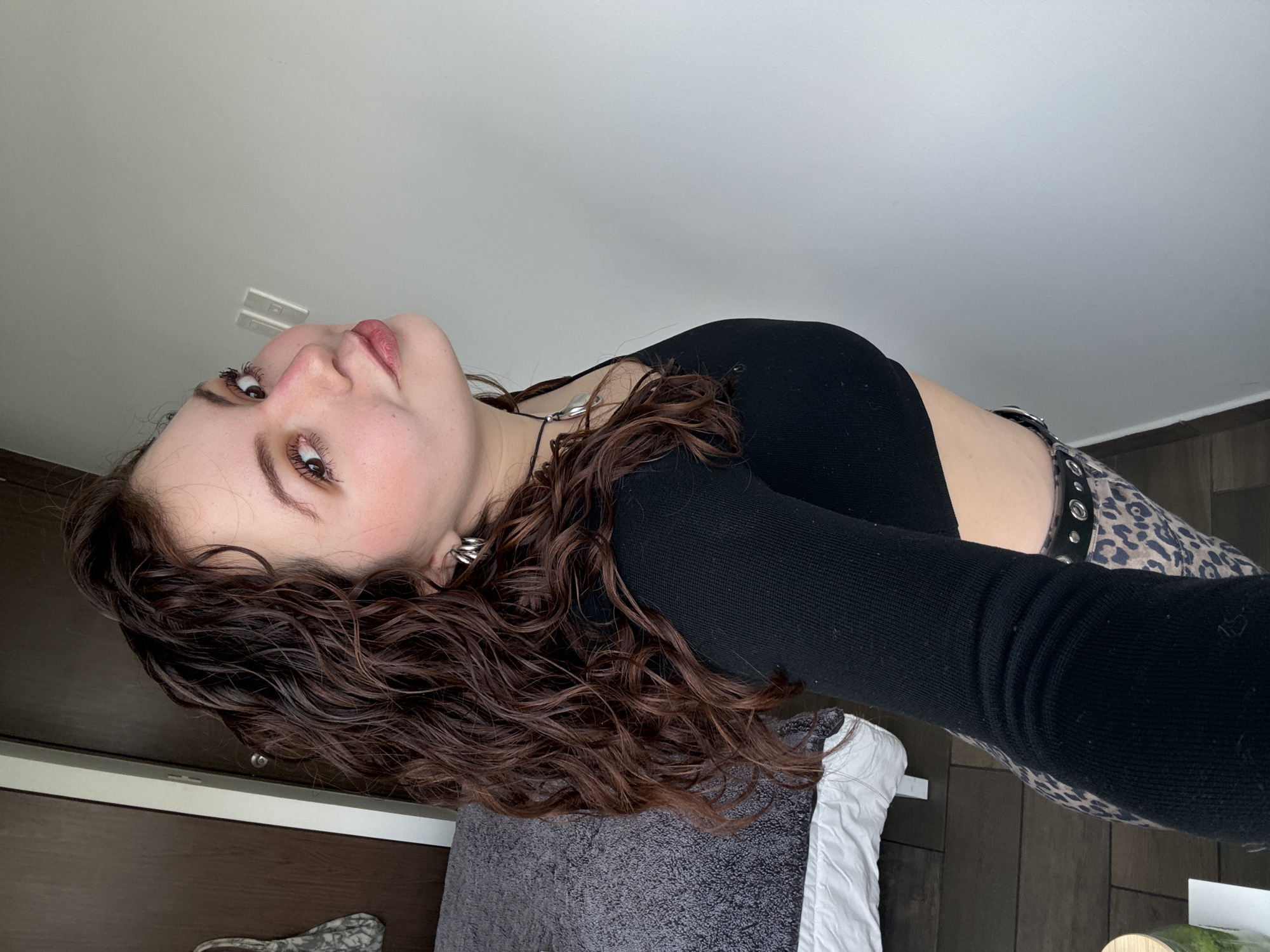 A young woman with light skin and brown, curly hair is depicted in a close-up, tilted portrait. She is wearing a black, long-sleeved crop top and animal-print pants with a studded belt. She has silver earrings and a necklace. She looks directly at the camera with a slight smile. The background includes a white ceiling with a light fixture, a dark wooden door with silver hardware, a gray textured cushion, and a wooden floor. The location is not explicitly indicated, and there are no elements to suggest that it is Mexico City.