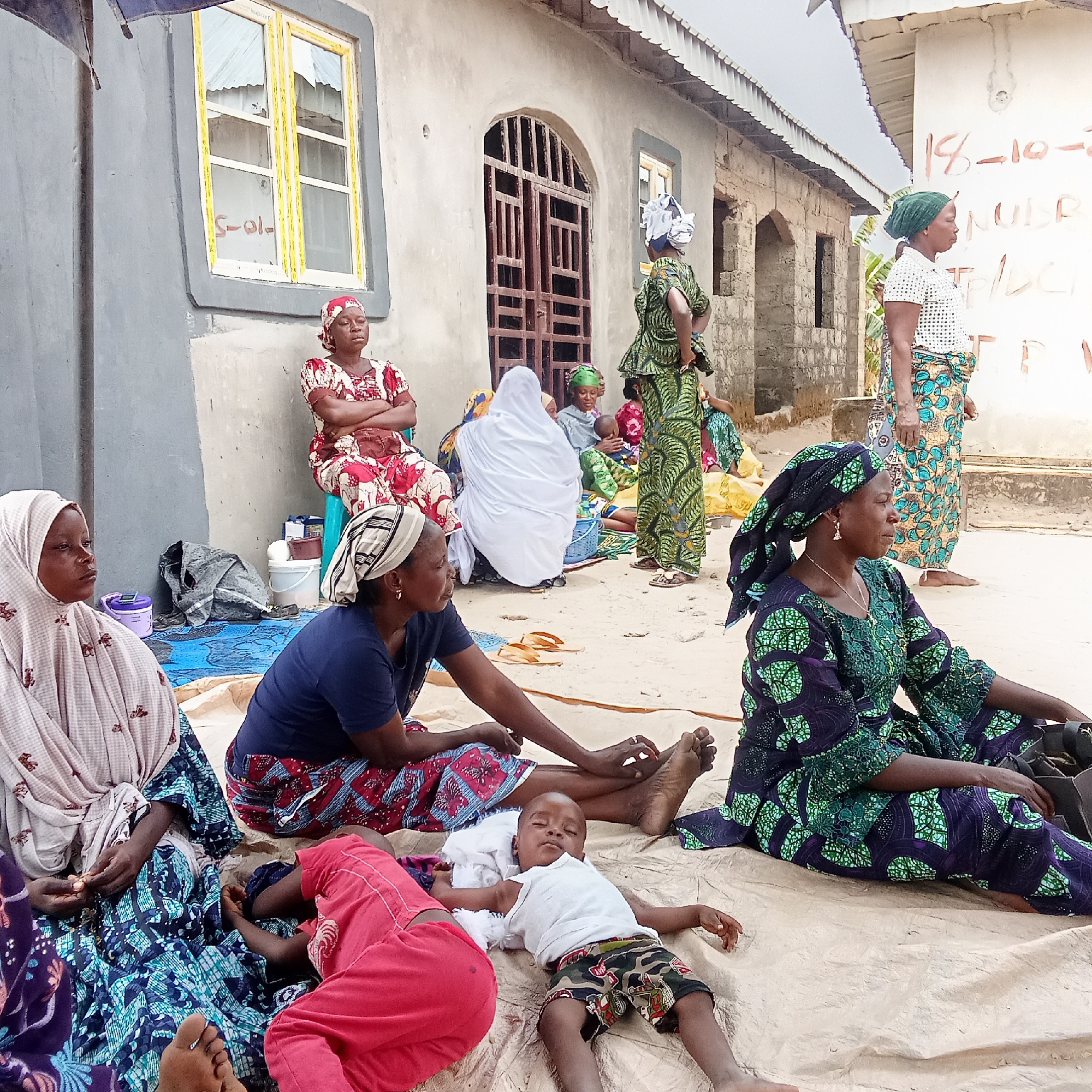 In Doma, Nigeria, a group of women are gathered outside a building. Some sit on mats on the ground, while others are standing or seated on a blue plastic stool. A child is lying on the mat. The women are dressed in patterned fabrics. The building has barred windows and some writing on the wall, including "18-10-2."