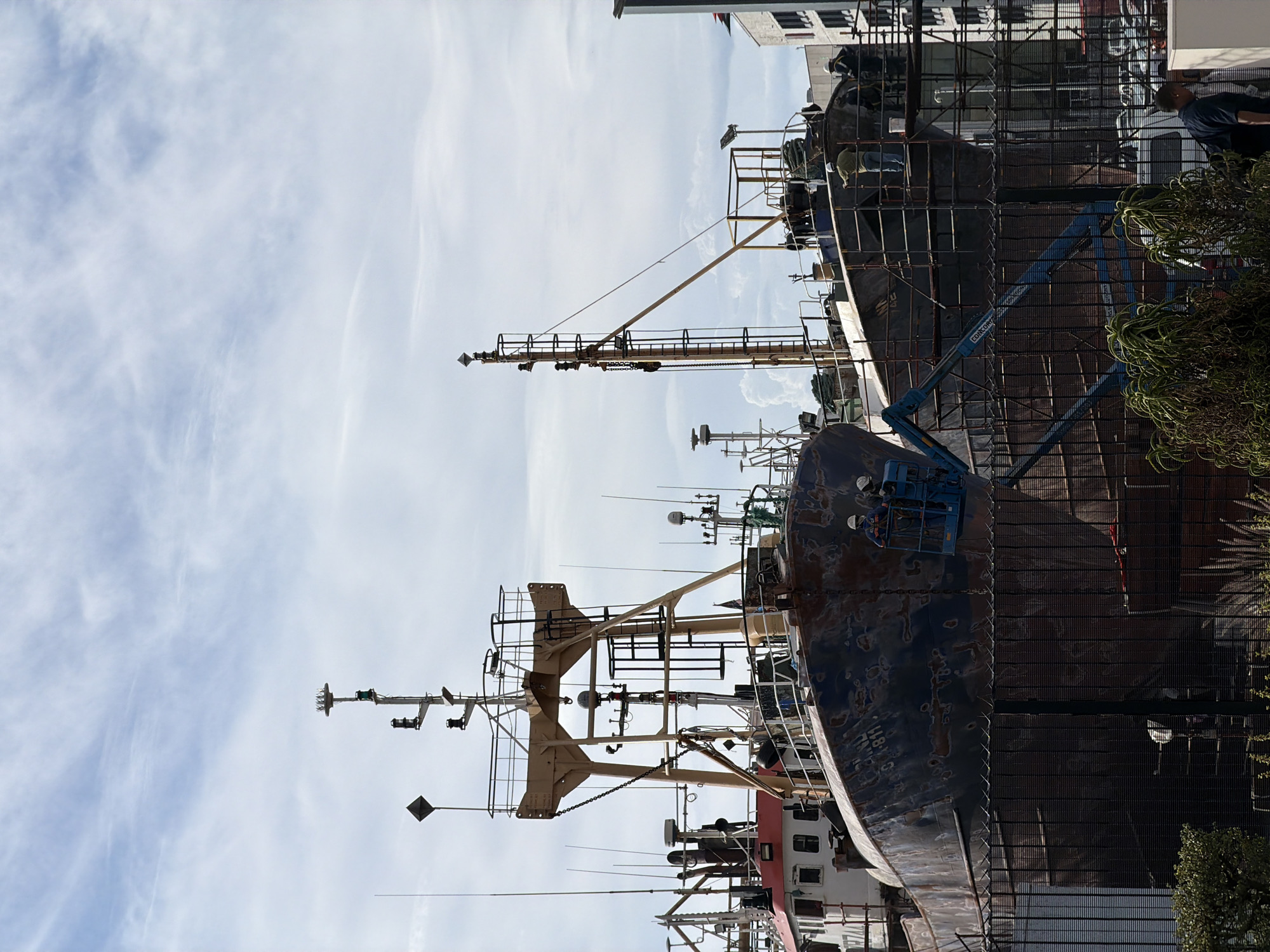 The image shows the side of a large fishing vessel under repair or renovation in Cape Town, South Africa. The vessel is dark blue and heavily rusted. Scaffolding surrounds the ship, and a blue cherry picker lift is visible near the bow. Two men are working on the upper part of the ship, wearing hard hats. A man stands on the dock, looking at the ship. A chain-link fence runs parallel to the vessel. Buildings in the background suggest the location is within an industrial or port area of Cape Town. The sky is light blue with wispy white clouds. A small bush sits in the right corner of the image.