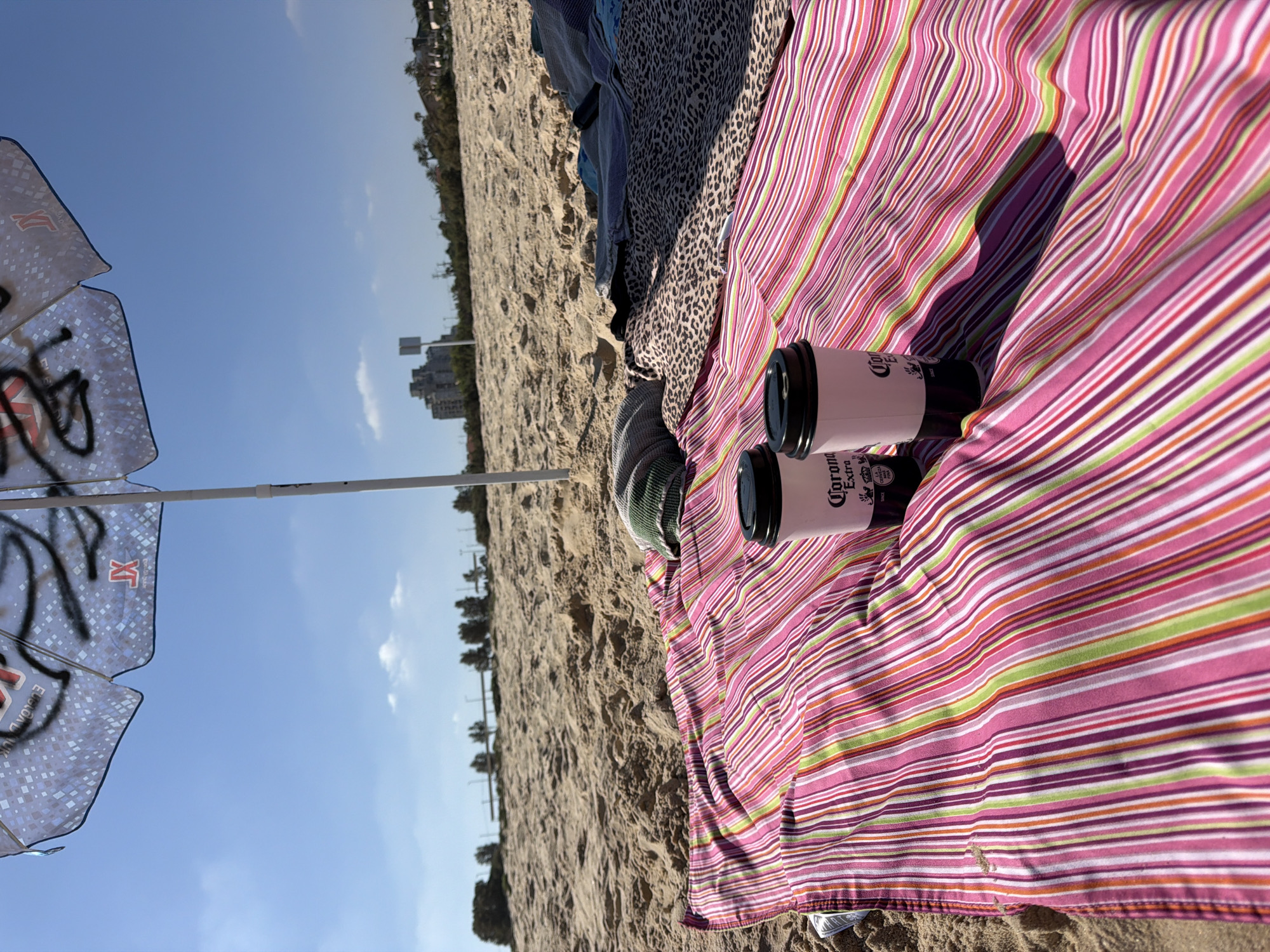 The image shows a beach scene in Ashkelon, Israel. A striped pink, white, green, and purple blanket is spread on the sand, partially covered by other blankets. Two white disposable cups with black lids, branded "Corona Extra", are placed on the blanket. An open beach umbrella, with a pattern of light blue squares and dark graffiti, stands behind the cups, casting a shadow. The background reveals a sandy beach, a line of trees and buildings in the distance, and a clear blue sky with a few white clouds. The overall impression is a relaxed, outdoor setting on a sunny day.