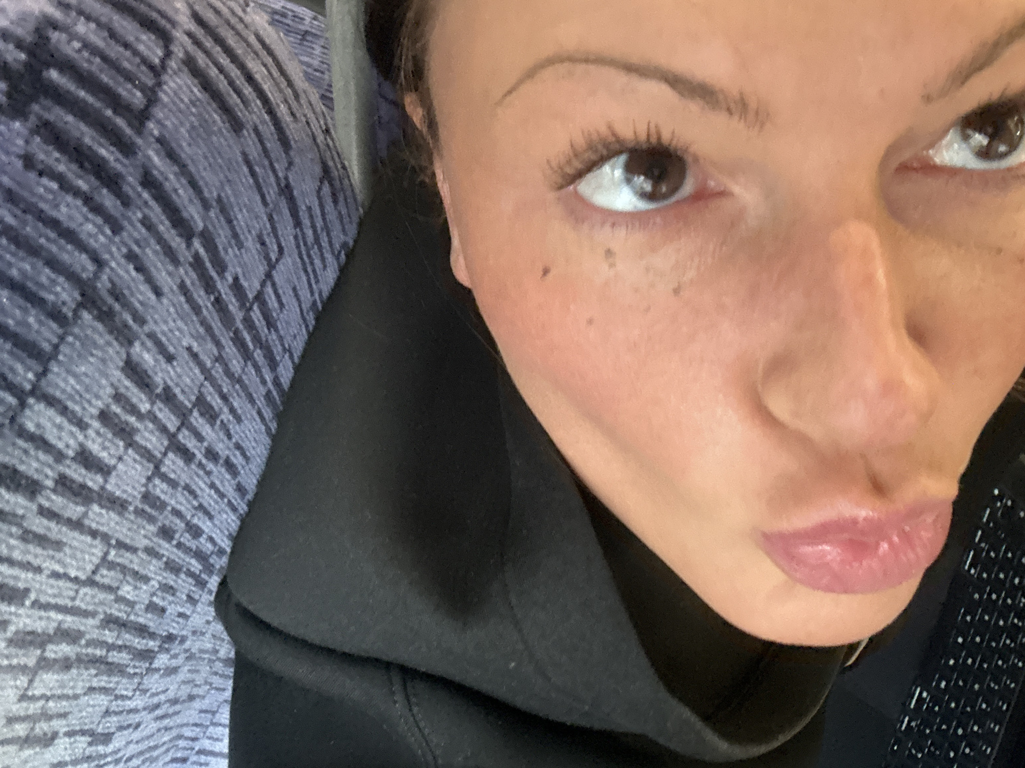 A selfie taken by a woman. She has brown eyes, thin eyebrows, and freckles on her face. She is pursing her lips. She is wearing a black hooded sweatshirt. To her left is a blue and grey patterned surface, possibly a seat. The location, Tunbridge Wells in the United Kingdom, cannot be confirmed from this image.