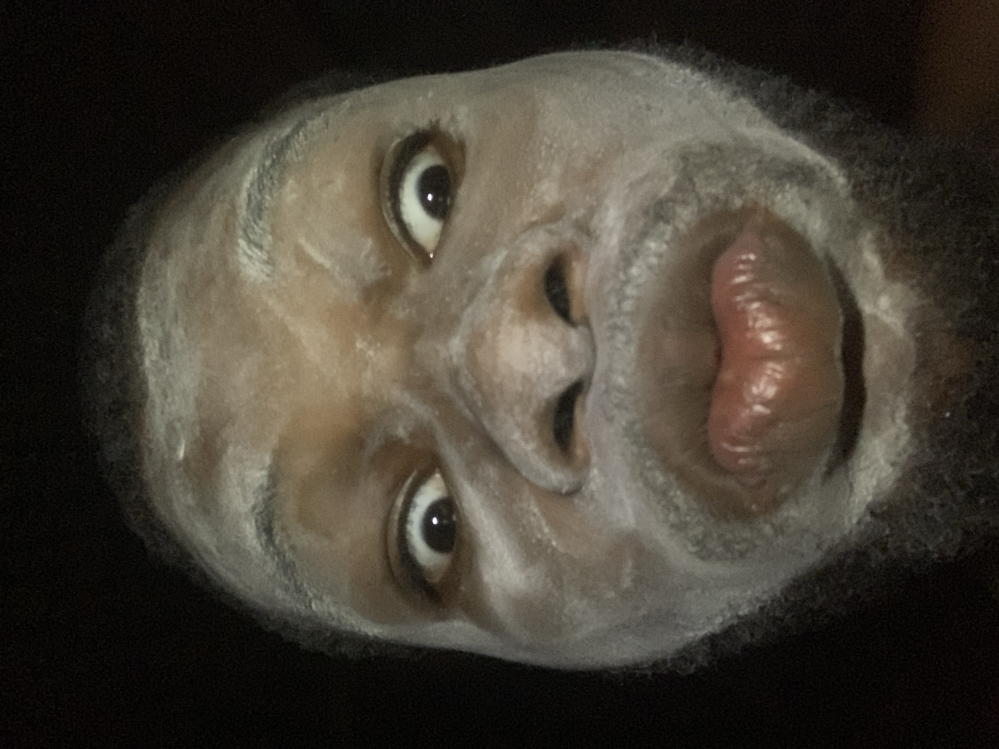 A close-up, rotated image shows the face of a dark-skinned man covered in a white powdery substance. The eyes are open, and the mouth is slightly open with a glossy appearance on the lips. The background is dark and indistinct. There is no verifiable information about the location being Onitsha, Nigeria, within the image itself.