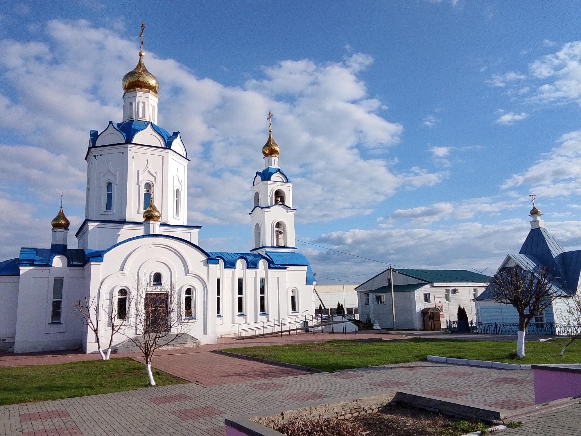 This daytime image showcases a vibrant Orthodox church complex under a bright blue sky dotted with scattered white clouds. The central feature is a large, white church building adorned with multiple golden onion domes, each crowned with a cross, and distinctive blue roofs. To the right of the main church is a prominent bell tower, similarly white with a blue roof and a golden dome and cross. Further to the right, a smaller, white building with a blue roof and another golden dome suggests a chapel or secondary religious structure.
In the background, a two-story white building with a green roof is visible, possibly a rectory or administrative facility, alongside other less defined structures. The foreground features well-maintained grounds with paved walkways made of reddish-brown and gray bricks, interspersed with patches of green grass. Several bare deciduous trees, some with their trunks painted white, are sparsely distributed across the landscape. The absence of leaves on the trees indicates either late autumn, winter, or early spring. No people are visible, and no specific activity is taking place. The architectural style is characteristic of Russian Orthodox churches, aligning with the location context of Khlevnoye, Russia. There is no visible text in the image.