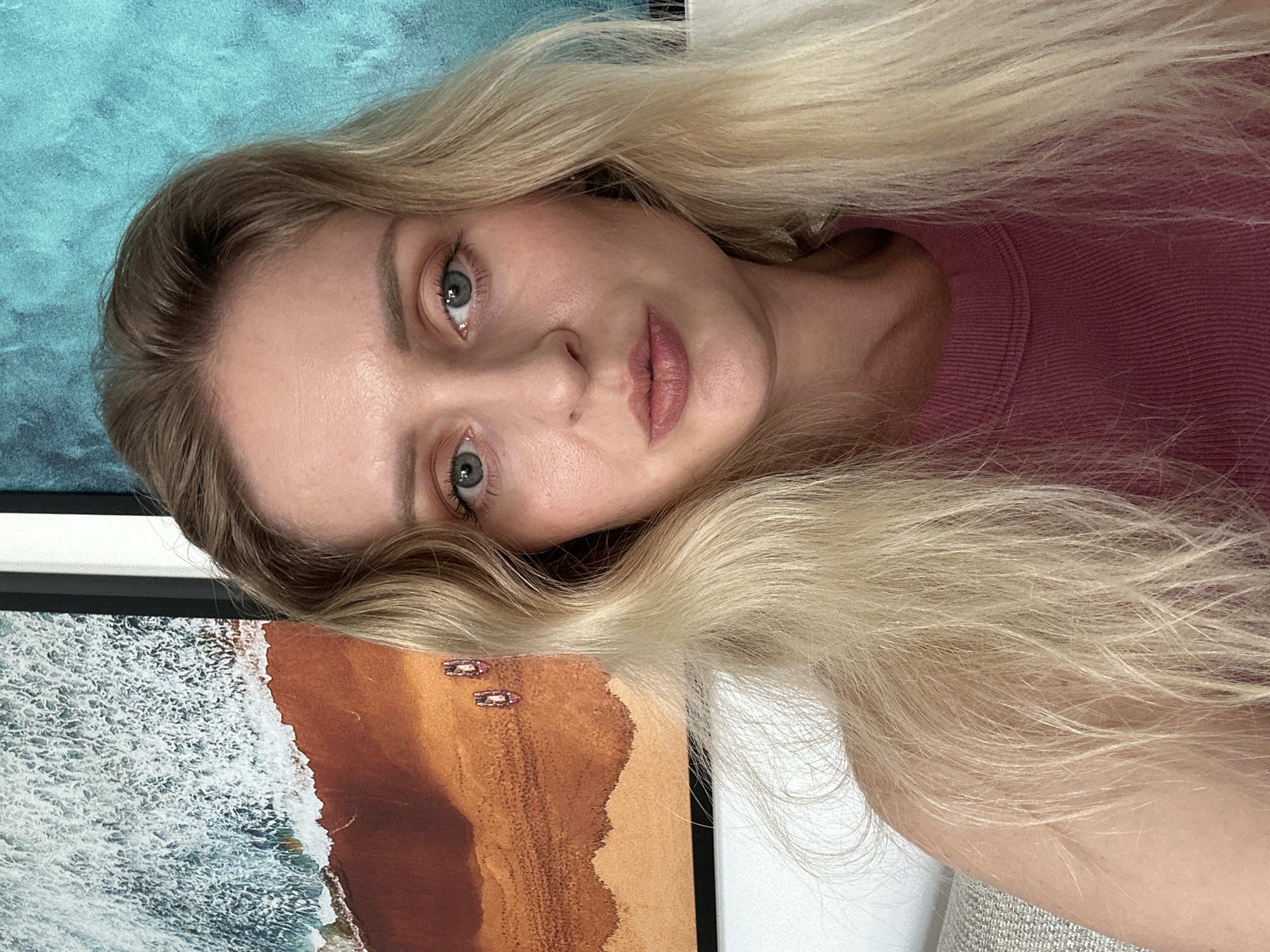 A fair-skinned woman with blonde hair and light blue eyes is in the foreground, wearing a rose-colored shirt. Behind the woman is an image of a beach with waves. There is no visible evidence of a specific location in Petah Tikva, Israel, nor are there any objects or interactions to suggest this setting.