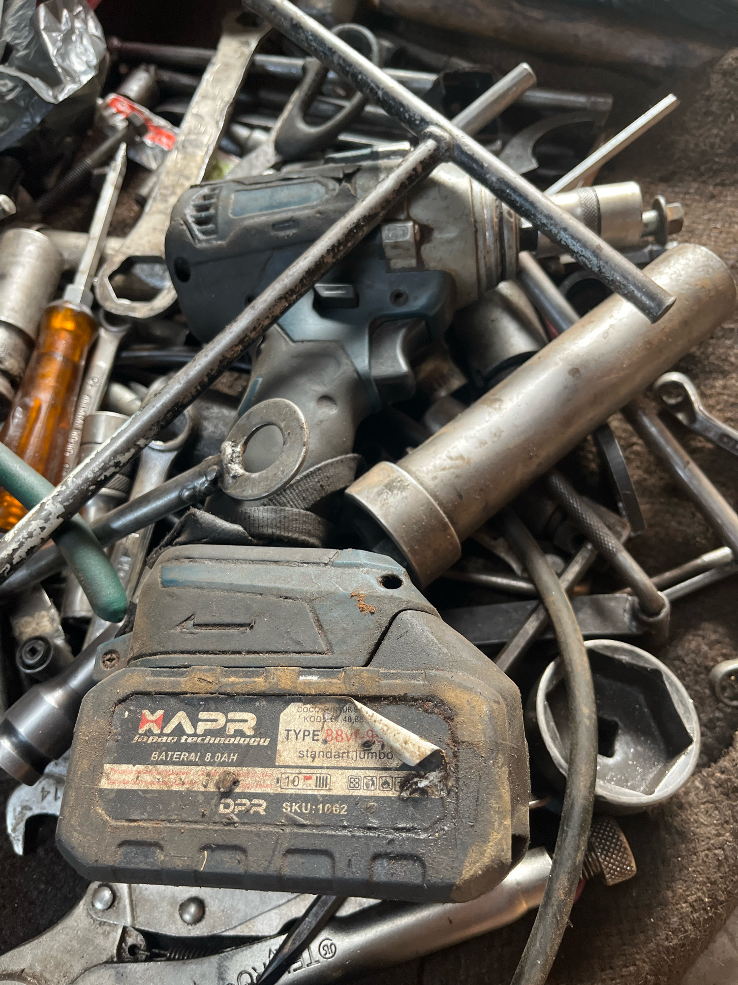The image shows a pile of used tools, including a cordless impact wrench and its battery pack, wrenches, sockets, and other hardware, resting on a brown, textured surface. The tools are dusty and appear to be in a workshop or storage area. The battery pack is labeled "XAPR JAPAN TECHNOLOGY," with text indicating "BATERAI 8.0AH," "TYPE BBV1-9," and "SKU:1062." There is no indication of the city of Kebumen, Indonesia, in the image or its contents.