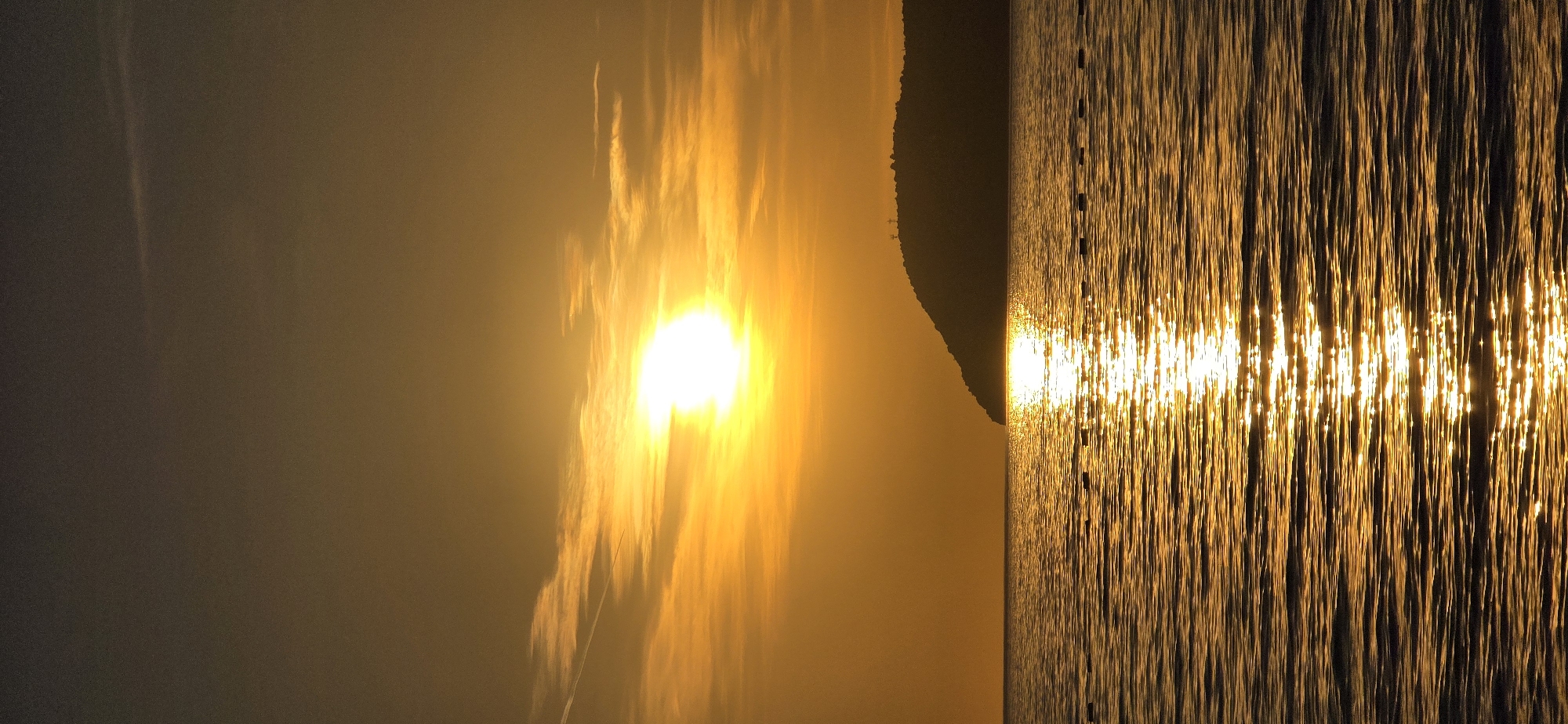 A vertical, long shot captures a sunrise or sunset over the Adriatic Sea in Bar, Montenegro. A bright sun shines through a hazy sky on the left side, creating long, shimmering reflections on the water to the right. A dark landmass breaks the horizon between the bright sky and the reflective sea.