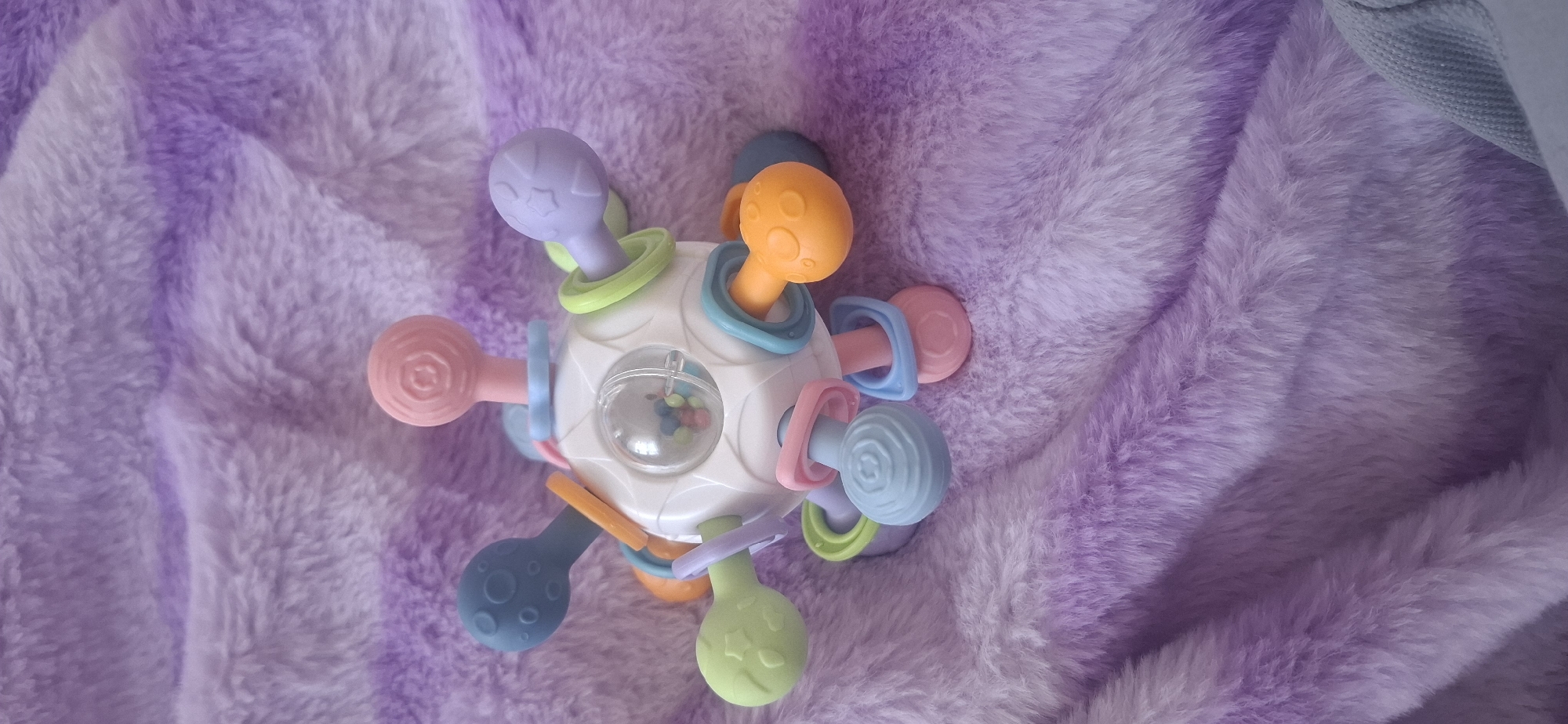 The image shows a colorful baby rattle toy with a clear spherical section containing small beads. The toy is placed on a soft, fluffy purple blanket. The toy has multiple flexible arms extending from a central white sphere. These arms are tipped with variously colored soft rubber shapes including circles, swirls, and textured balls in pink, blue, green, purple, and orange. The blanket exhibits a gradient of light to dark purple hues and appears to be made of a plush material. No people are present in the image. The location of Ust'-Katav, Russia is not depicted or suggested by any elements within the image.