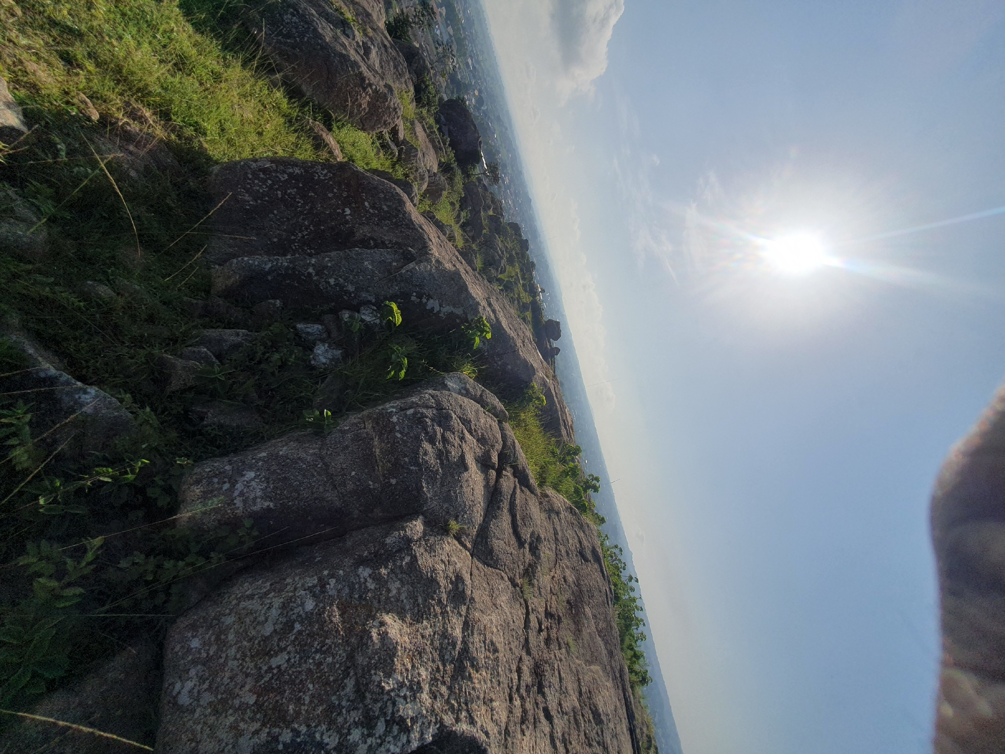 The image shows a view from a rocky outcrop overlooking a city, identified as Gura Top, Nigeria. The foreground is dominated by large, weathered rocks covered in sparse green vegetation. The rocks are rough and uneven, with visible cracks and lichens. In the midground, a sprawling city is visible below, with houses and buildings spread across the landscape. The background consists of a hazy horizon with distant hills. The sky is bright blue with a few scattered clouds, and the sun is shining, creating a lens flare effect in the upper left quadrant of the image. A blurry, dark object, possibly a hand or part of a camera, is visible in the extreme upper portion of the image, suggesting the photo was taken from a handheld perspective. The overall scene depicts a natural landscape juxtaposed with an urban environment under a bright, sunny sky.