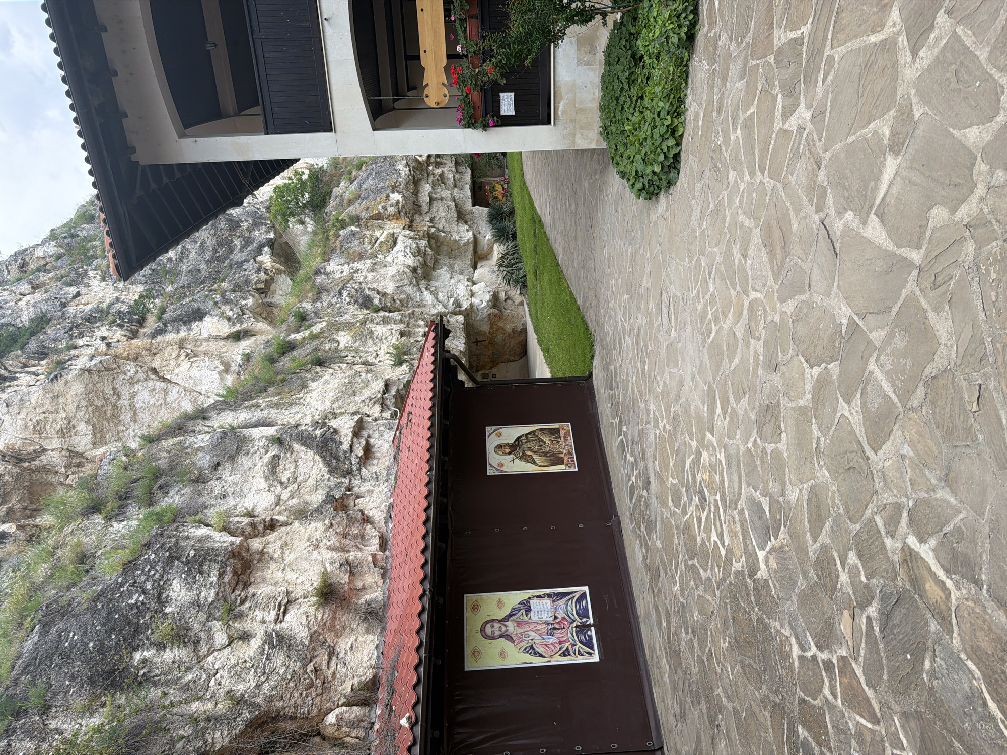 This image depicts a section of the Basarbovo Monastery in Basarbovo, Bulgaria, situated at the base of a significant rock formation. The scene is dominated by a rugged, light-colored limestone cliff face that rises sharply in the background, showing varying textures, shadows, and sparse green vegetation growing from crevices.
In the foreground, a flagstone-paved courtyard extends across the lower portion of the image. The paving consists of irregularly shaped, flat grey and brown stones set with light-colored mortar.
On the left side of the courtyard, a low, rectangular structure with a red, ridged tiled roof and a dark brown, curtain-like facade is visible. Two large rectangular religious icons are affixed to this facade: the icon on the left portrays Jesus Christ holding a book, while the icon on the right depicts a monastic figure in dark robes, possibly Saint Dimitar Basarbovski, holding a staff.
To the right of the paved area, a manicured strip of green grass and foliage separates the foreground from a pathway leading deeper into the monastery grounds, towards additional cave openings visible in the cliff face.
On the far right, a portion of a multi-story building is visible. This structure features light-colored, possibly beige or white, plastered walls and a dark tiled roof with exposed wooden beams beneath the eaves. An arched opening reveals a dark-railed balcony on the upper floor. On the lower visible section, a wooden sign or hanging element is suspended, and a dark window or door frame is adorned with a window box containing vibrant red and pink potted flowers. A small, leafy green bush grows in the corner of the courtyard near this building.
The sky above is overcast, providing diffuse lighting for the scene. No individuals are present in the photograph.
