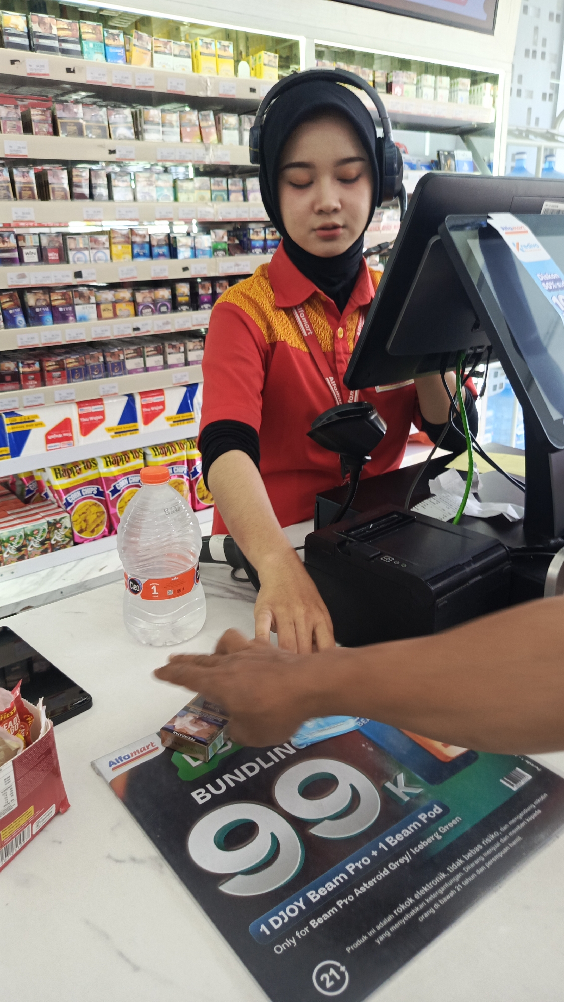 In an indoor convenience store, likely an Alfamart branch in Tangerang, Indonesia, a young female cashier wearing a red and yellow uniform and a black hijab is attending to a customer. She has headphones on and appears to be focused on the transaction. A customer's hand is visible, reaching towards a pack of cigarettes placed on a promotional flyer with the text "BUNDLING 99K" and "1 DJOY Beam Pro + 1 Beam Pod". Behind the cashier, shelves are stocked with various products, including snacks and cigarette packs. A large bottle of water is on the counter in front of her. The scene depicts a typical retail interaction at a convenience store checkout.