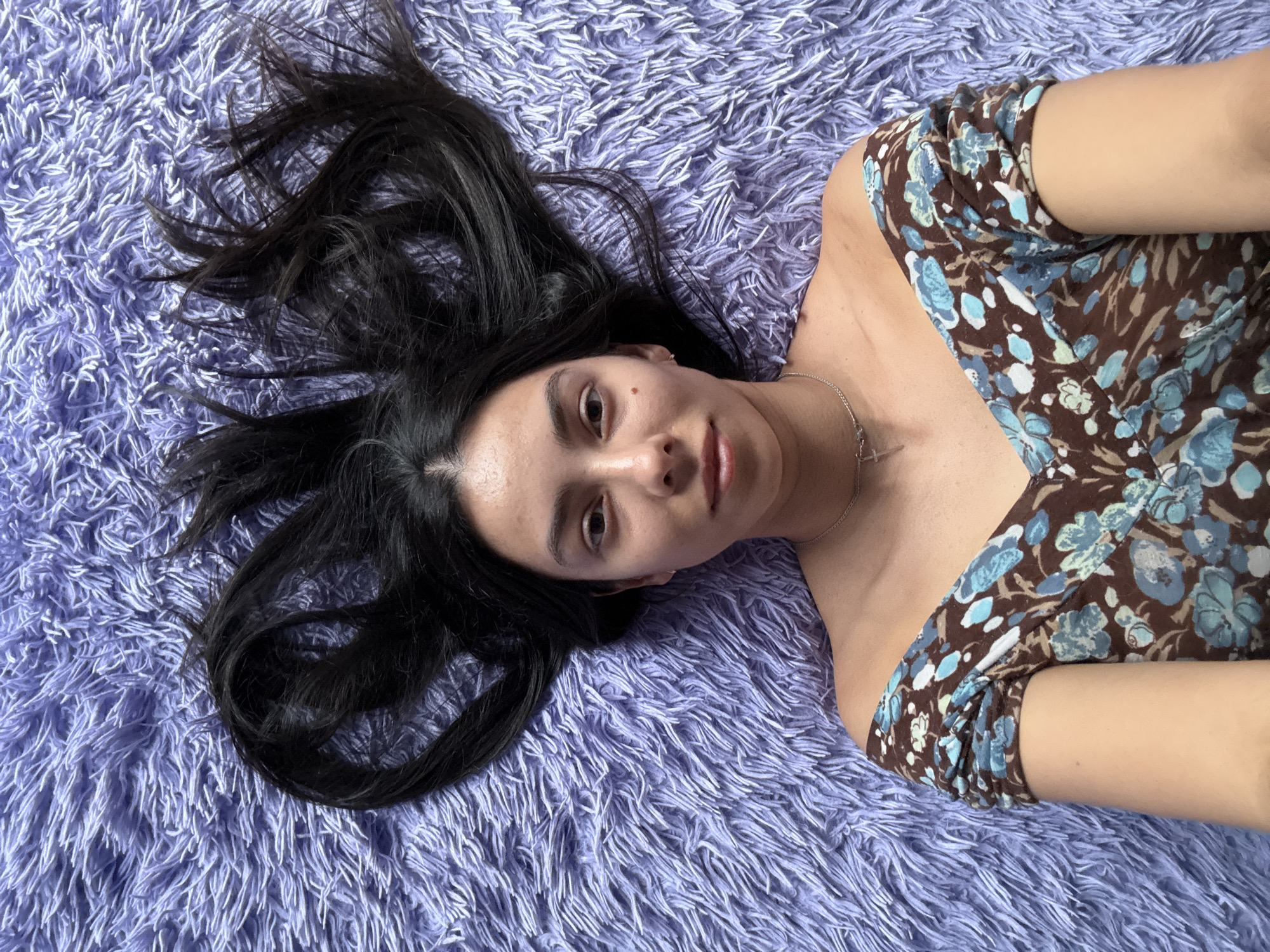 A young woman lies on a purple shag carpet. She has long, dark hair fanned out on the carpet. She is wearing a brown floral-print top with blue flowers. She has a silver chain with a cross pendant around her neck. The location is not apparent from the image and cannot be determined to be in Chaikovsky, Russia.