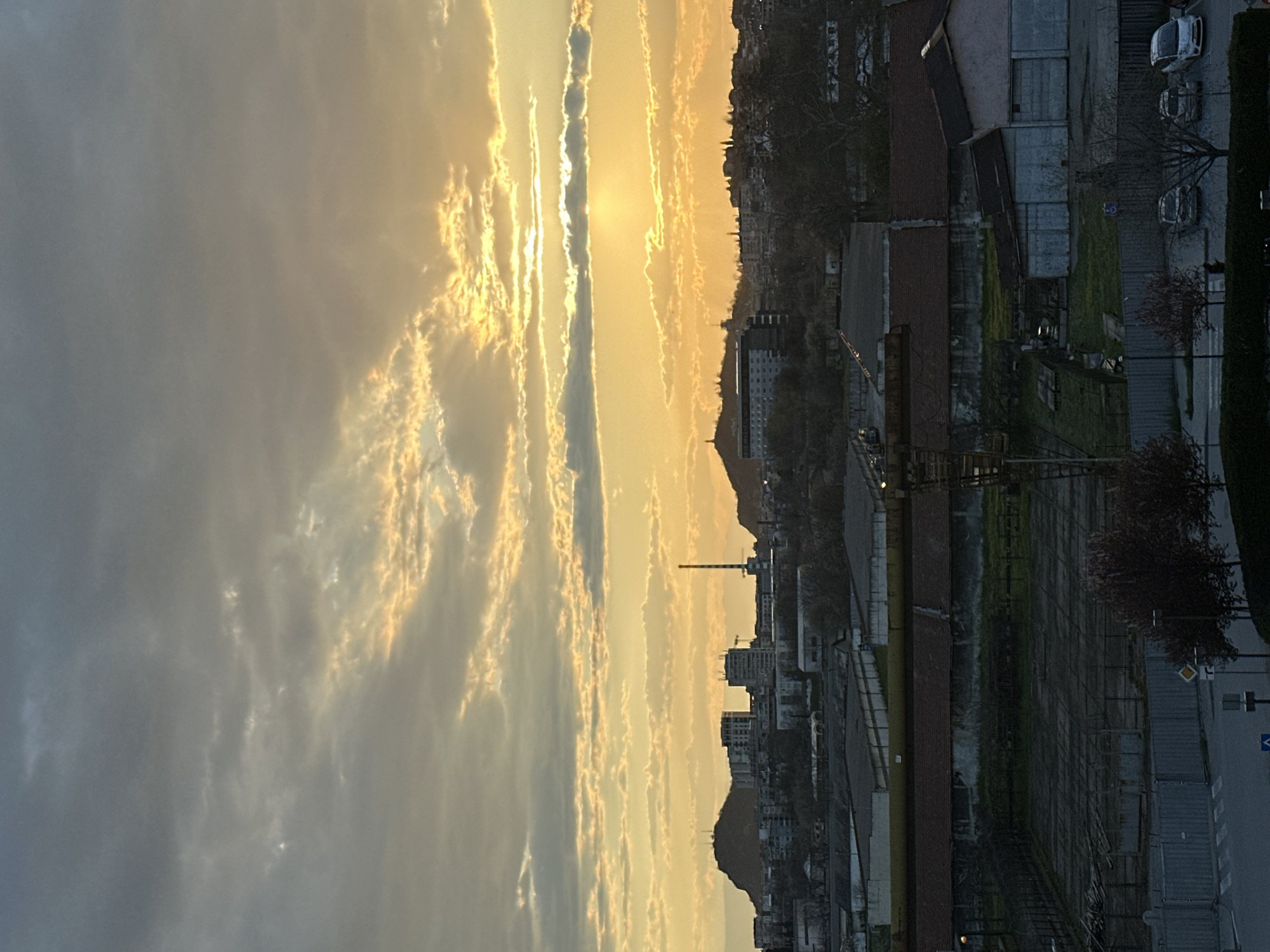 The image captures a cityscape during what appears to be a sunset in Plovdiv, Bulgaria. A sky filled with striated clouds is illuminated by the setting sun. The built environment is dominated by warehouses, buildings, trees, and streets.
A prominent feature is the Maritsa River. It is visible on the right side of the frame along with warehouses. The overall tonality of the city is muted by the light, suggesting an industrial district.