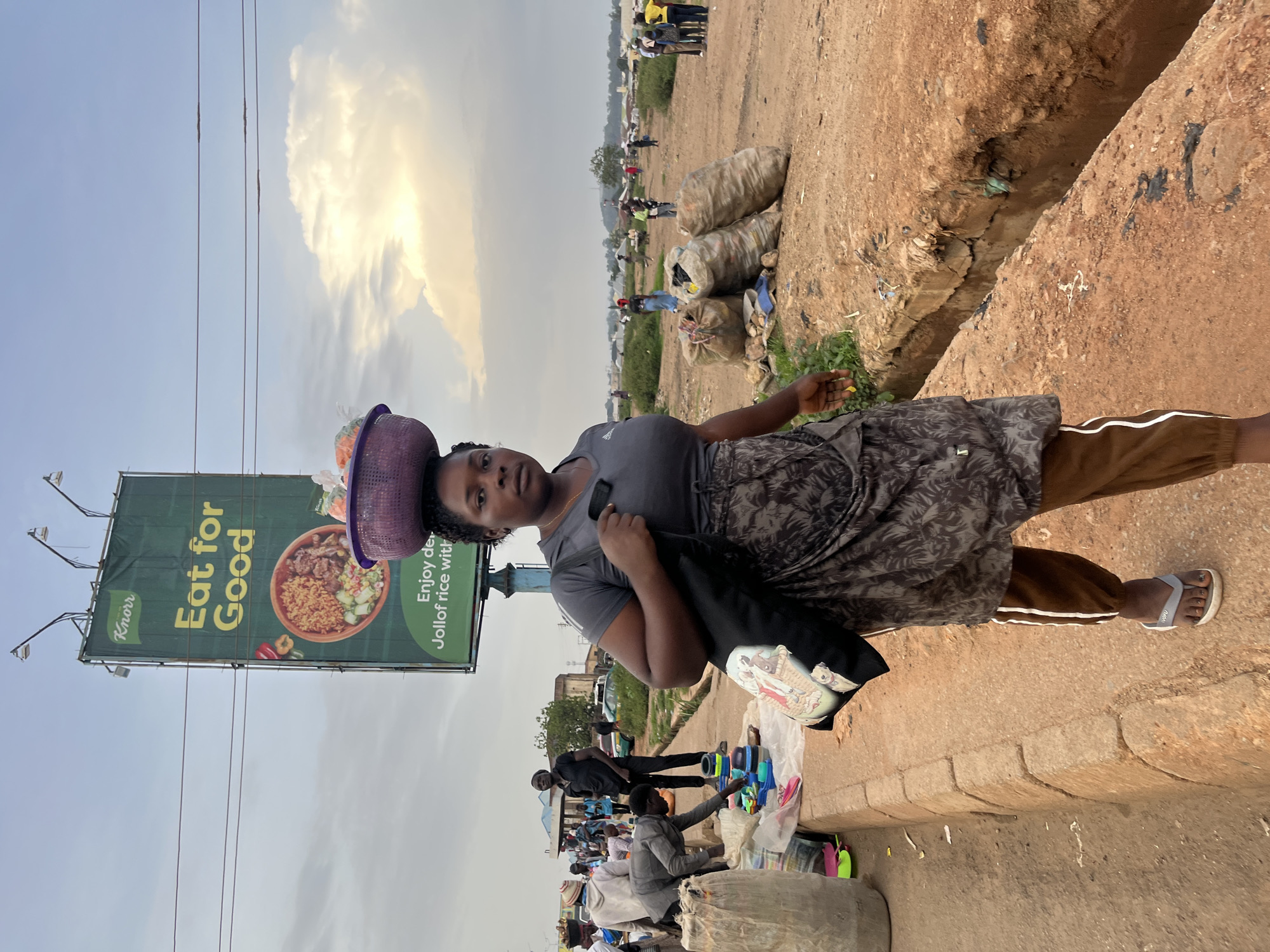 A woman stands on a dirt road in Bukuru, Nigeria, carrying a basket of items on her head. She is wearing a dark gray t-shirt, a patterned skirt, and brown pants with white stripes. A black bag hangs from her shoulder, featuring an illustration of a person. The scene appears to be a market or roadside stall area, with other people and merchandise visible in the background. Piles of sacks are stacked near the road, and a ditch runs alongside it. A large billboard for Knorr, advertising "Eat for Good" and "Enjoy delicious Jollof rice," is prominently displayed. The sky is cloudy with sunlight breaking through, suggesting it is either early morning or late afternoon. The overall impression is of a bustling outdoor environment, possibly a commercial hub in Nigeria.