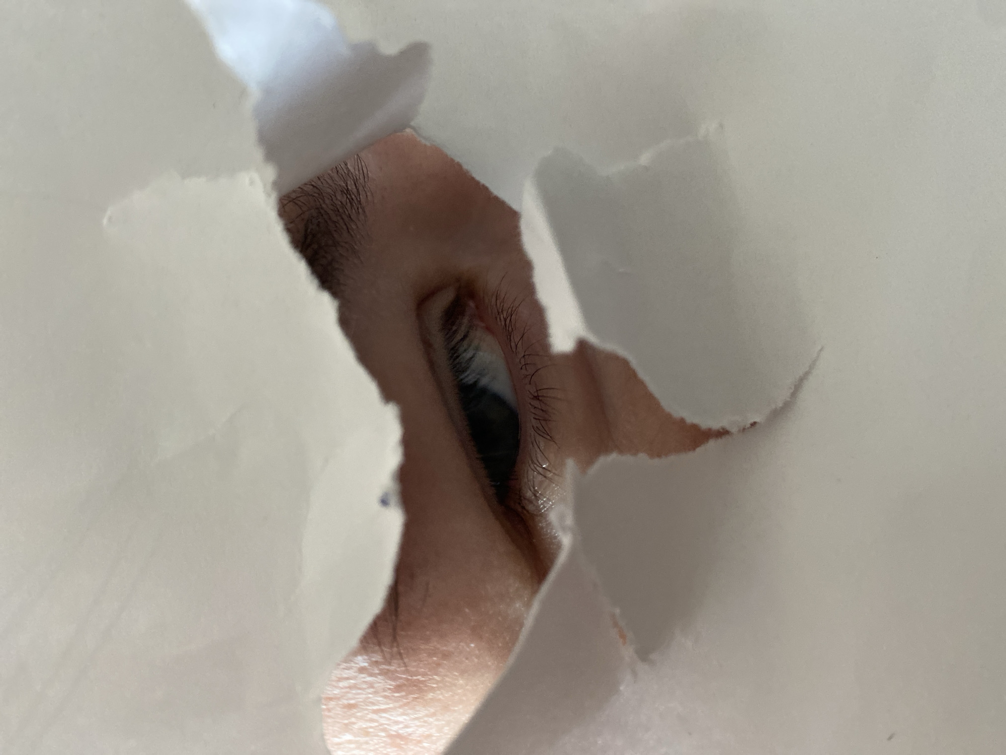 The image features a close-up of a human eye peering through a jagged, irregular opening in what appears to be white or off-white paper or fabric. The eye is positioned at an upward and slightly rightward angle relative to the torn edges. The skin around the eye, including the eyelid, is visible, along with distinct eyelashes and a portion of an eyebrow. The iris of the eye appears dark. The edges of the torn material create a natural, rough frame around the eye, emphasizing the act of looking through a small aperture. The lighting highlights the texture of the torn material and the skin around the eye. No other subjects, activities, or text are discernible in the tightly cropped frame. The scene suggests an act of clandestine observation, peeking, or surveillance.