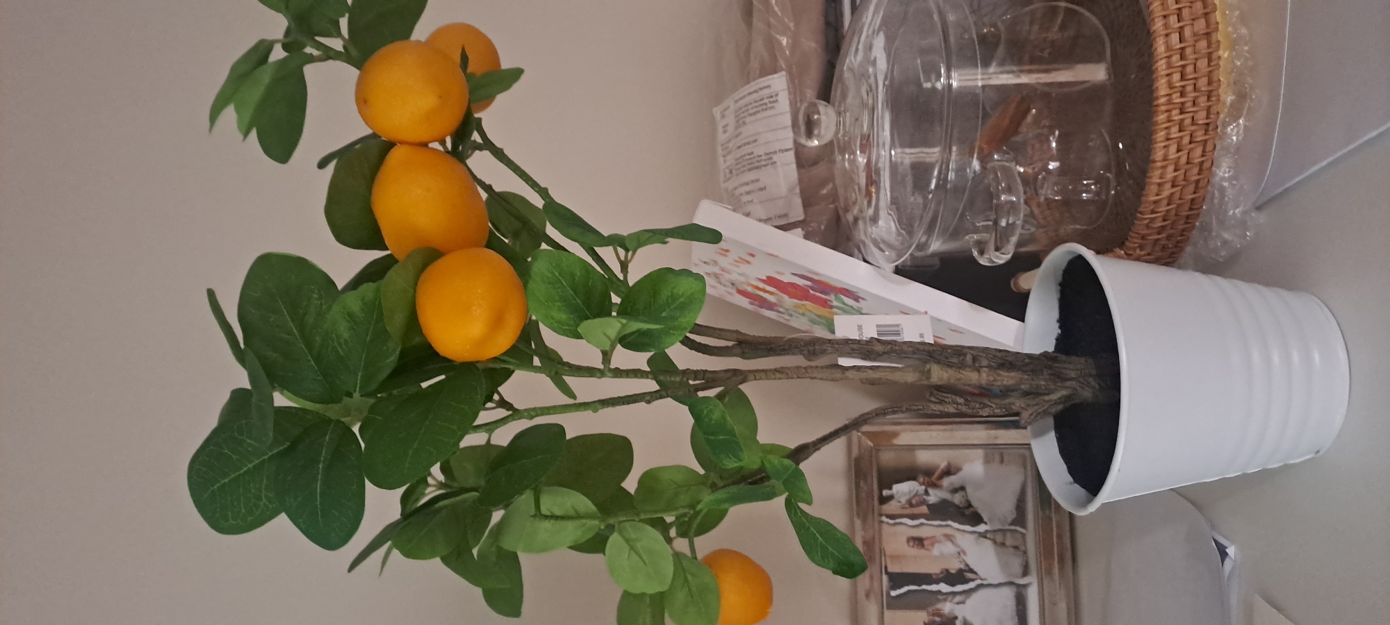 The image shows a close-up of a faux lemon tree in a white pot, positioned on a shelf. The tree has green leaves and several yellow artificial lemons. Behind the tree, to the left, is a framed photograph of a wedding couple. To the right of the tree, there is a stack of books or a notepad with floral illustrations, and behind that, a clear glass jar with a lid. The jar appears to contain some items, possibly cinnamon sticks or similar. The background is a plain, light-colored wall. There are no people visible in the image. The scene appears to be indoors, possibly in a home or retail setting. The location of Castlebar, Ireland is not depicted in the image.