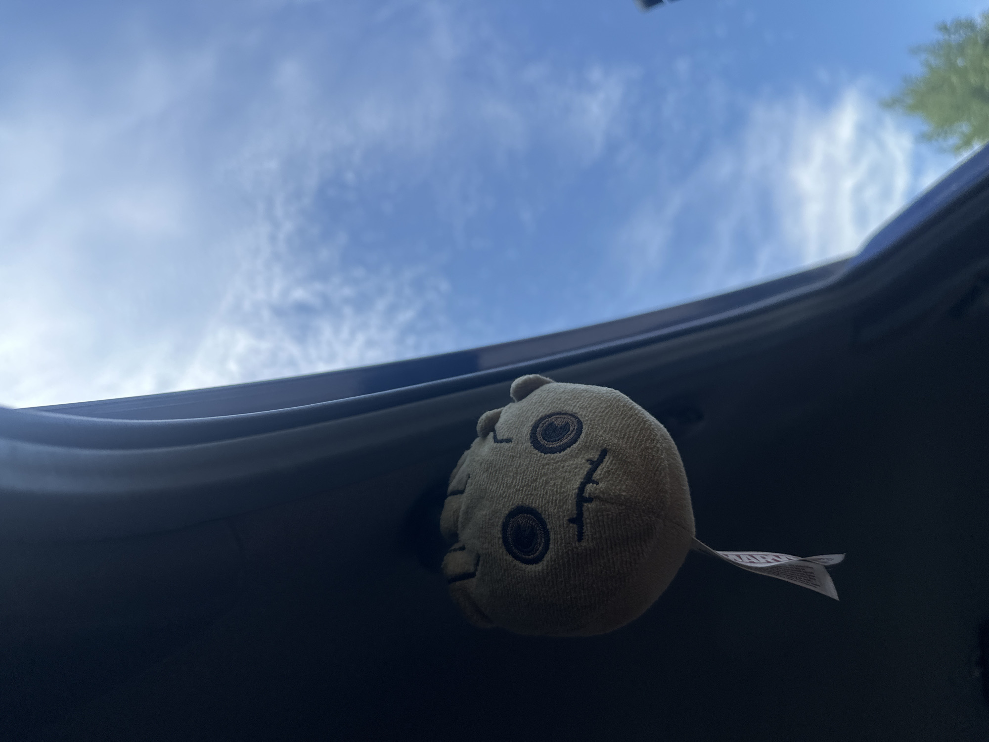 A plush toy resembling Groot is attached to the interior of a vehicle, possibly a car trunk or hatchback, viewed from inside the vehicle looking upwards. The plush toy, which is light brown with embroidered dark eyes and a mouth, hangs from a white tag. The background is a bright blue sky with scattered white clouds, and a hint of green foliage is visible in the lower right corner. The lighting suggests it is daytime. The location is not specified in the image or its context.