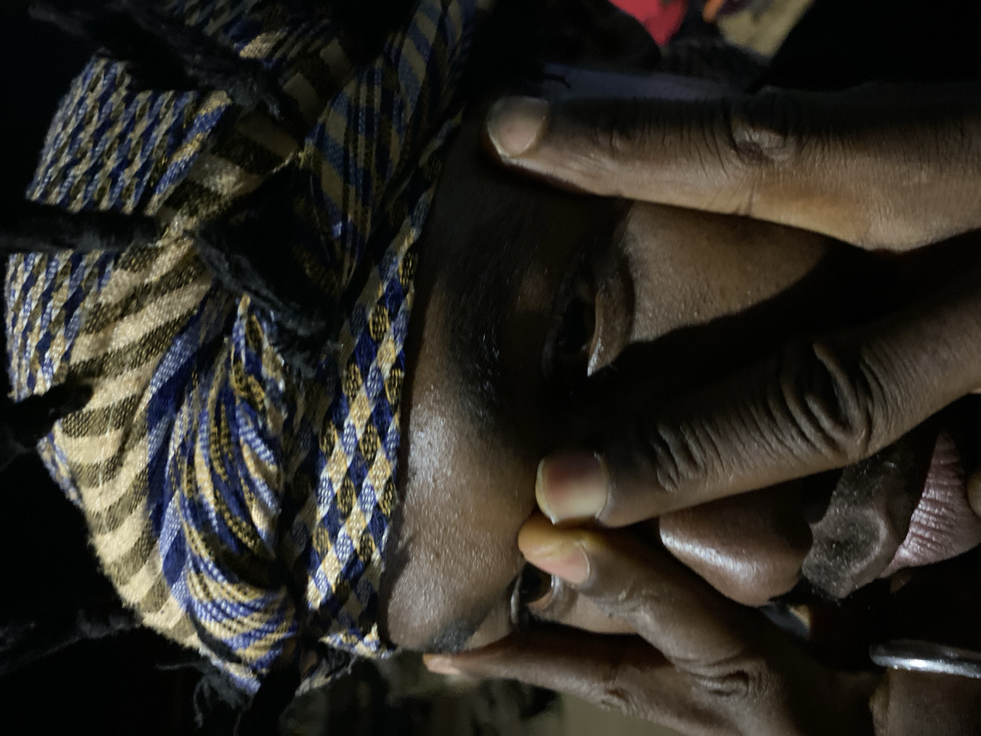 A close-up, low-angle, outdoor shot captures a person wearing a checkered headscarf of blue, yellow, and beige stripes. The individual's face is partially obscured by their hands, which are positioned near their eyes and nose. One hand, with fingers extended, covers the left side of their face, while the other hand, with fingers bent, is near the right eye. The person has dark skin and visible stubble on their upper lip and chin. A silver ring is visible on the left index finger. The lighting is dim, creating strong shadows and highlighting certain features of the face. The background is dark and out of focus, with hints of color that might suggest clothing or other objects. The photograph appears to have been taken in Bukuru, Nigeria, based on the provided context.